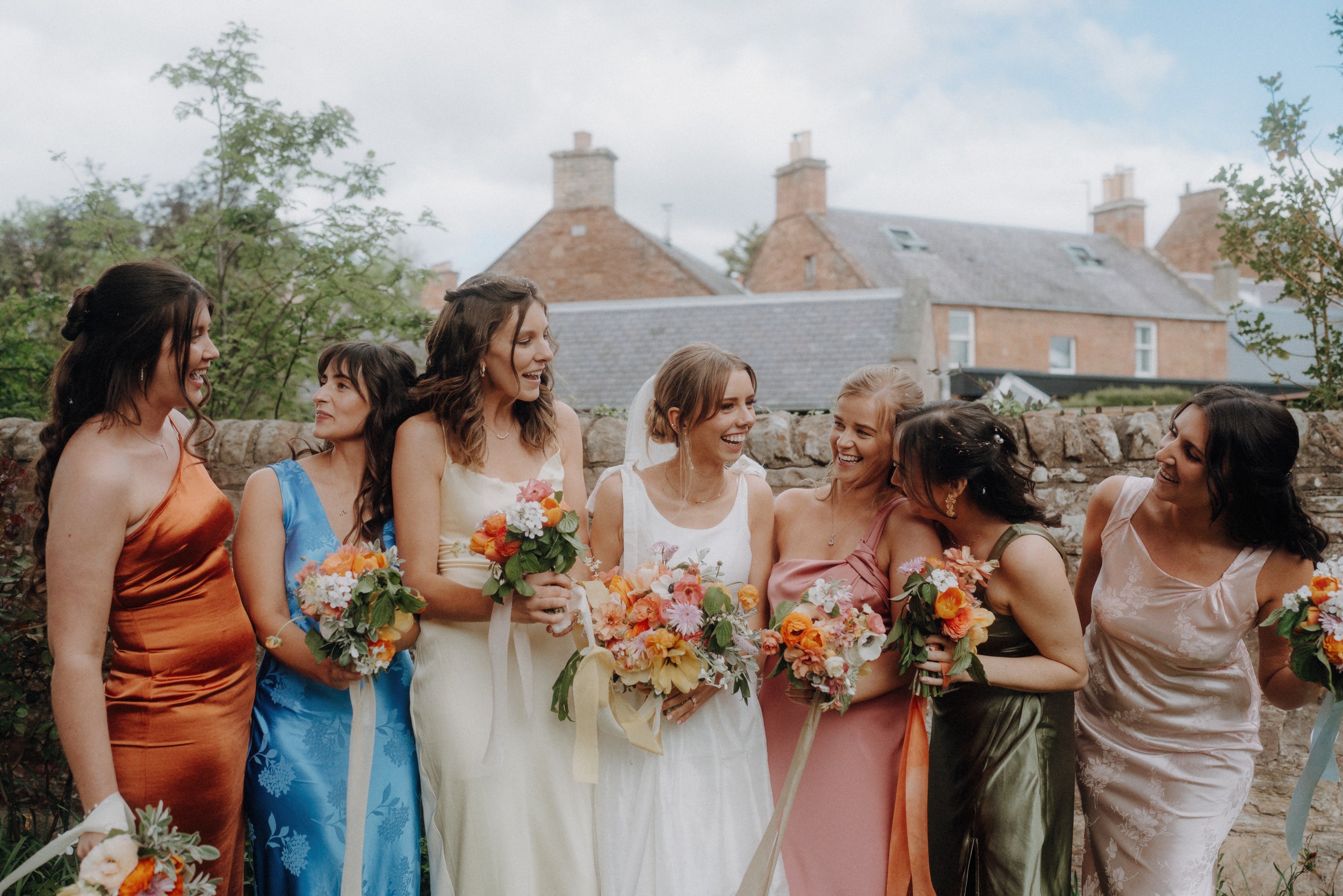 Seven women in colorful dresses stand outdoors holding bouquets, smiling and laughing together, with stone walls and houses in the background.