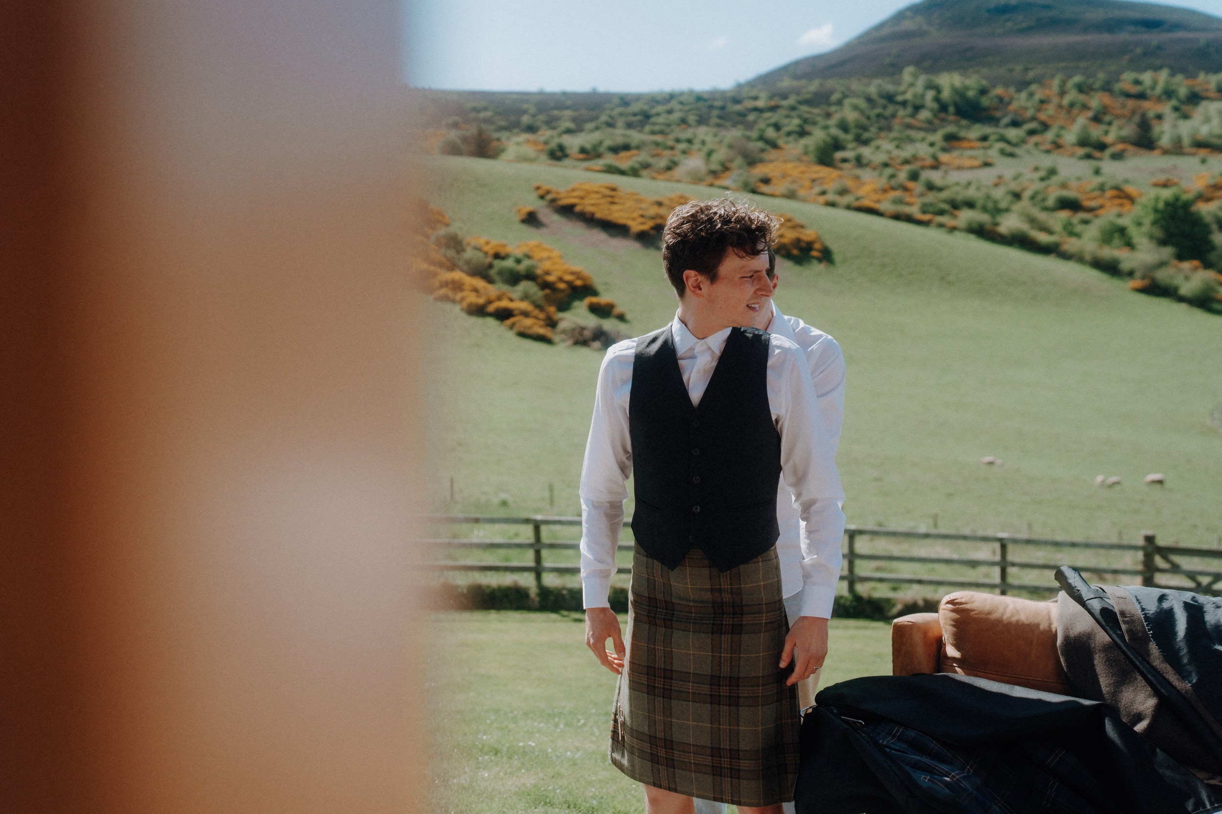A man wearing a vest and a kilt stands outdoors on a grassy hillside with sheep and yellow-flowering bushes in the background.