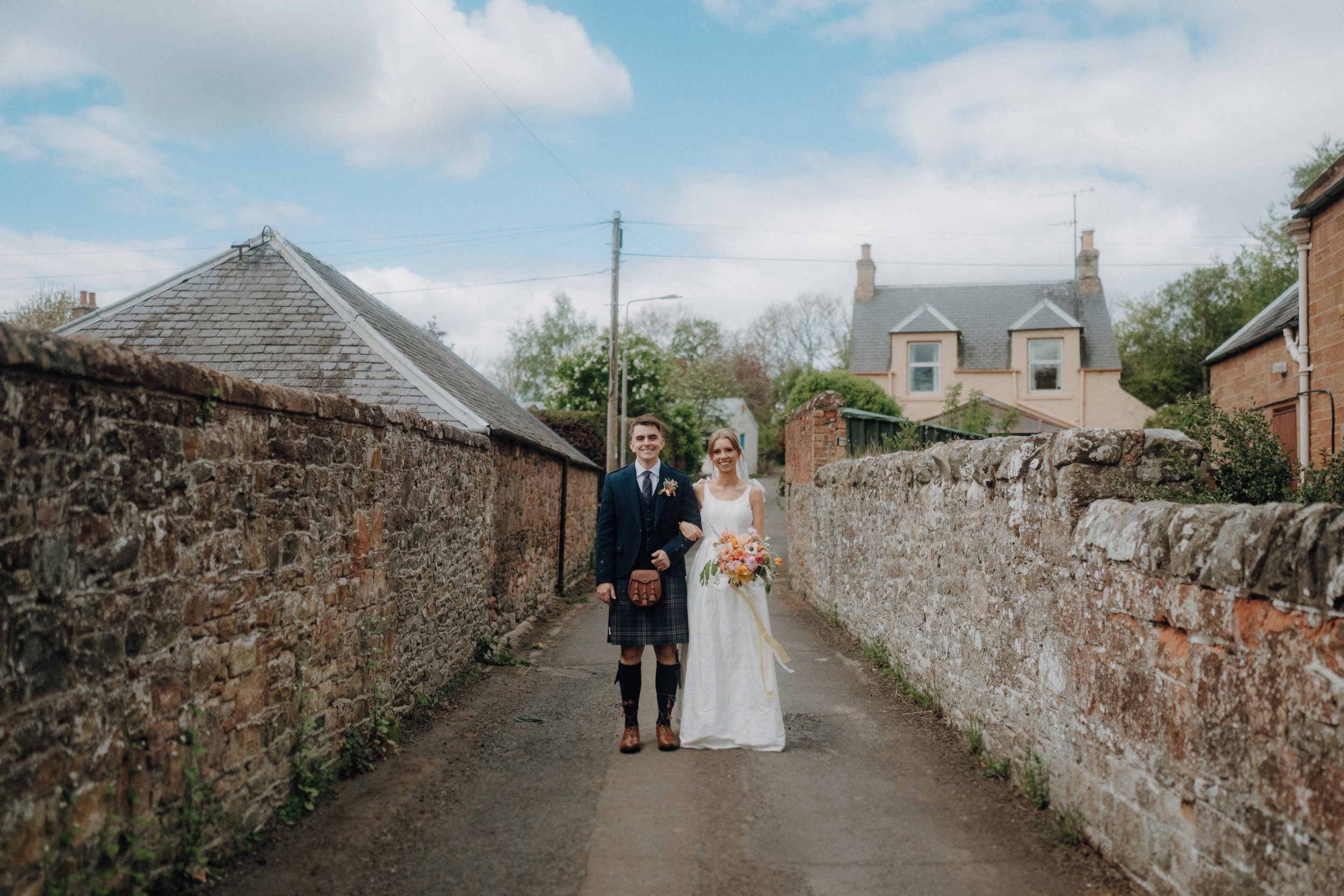 A bride and groom stand side by side in wedding attire on a narrow lane between stone walls, with houses and greenery in the background.