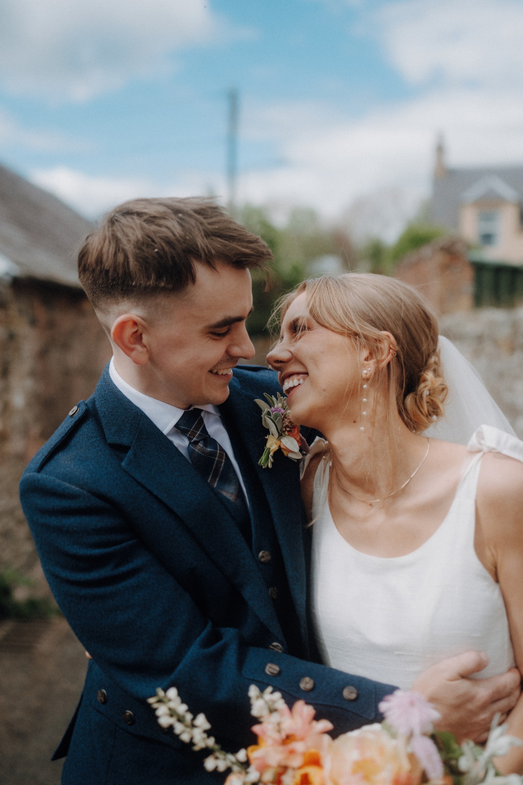 A newly married couple dressed in formal attire smile and embrace outdoors, with the bride holding a bouquet and the groom in a blue suit.