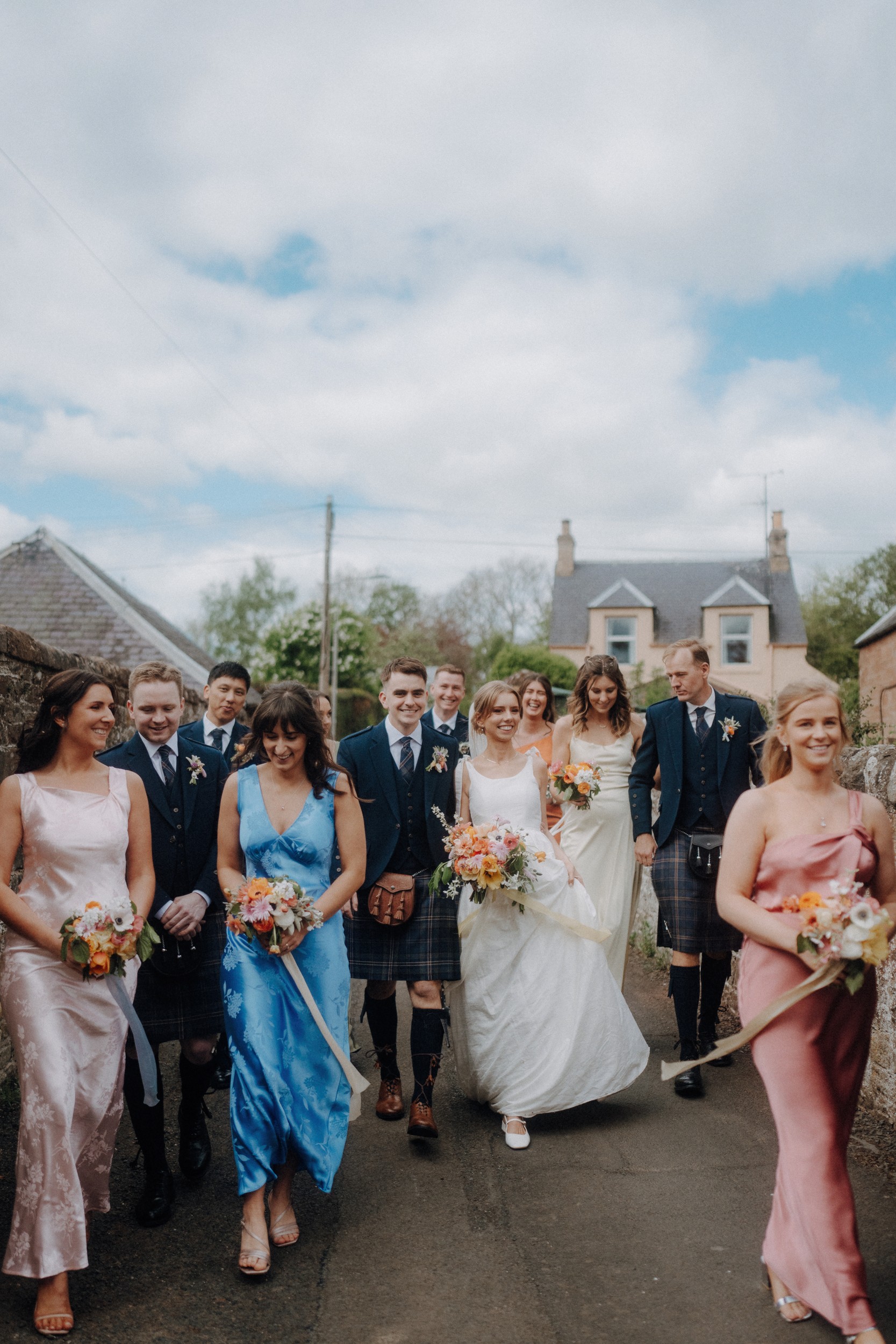 A wedding party in formal attire, with bridesmaids in colorful dresses and groomsmen in kilts, walk down an outdoor path holding bouquets.
