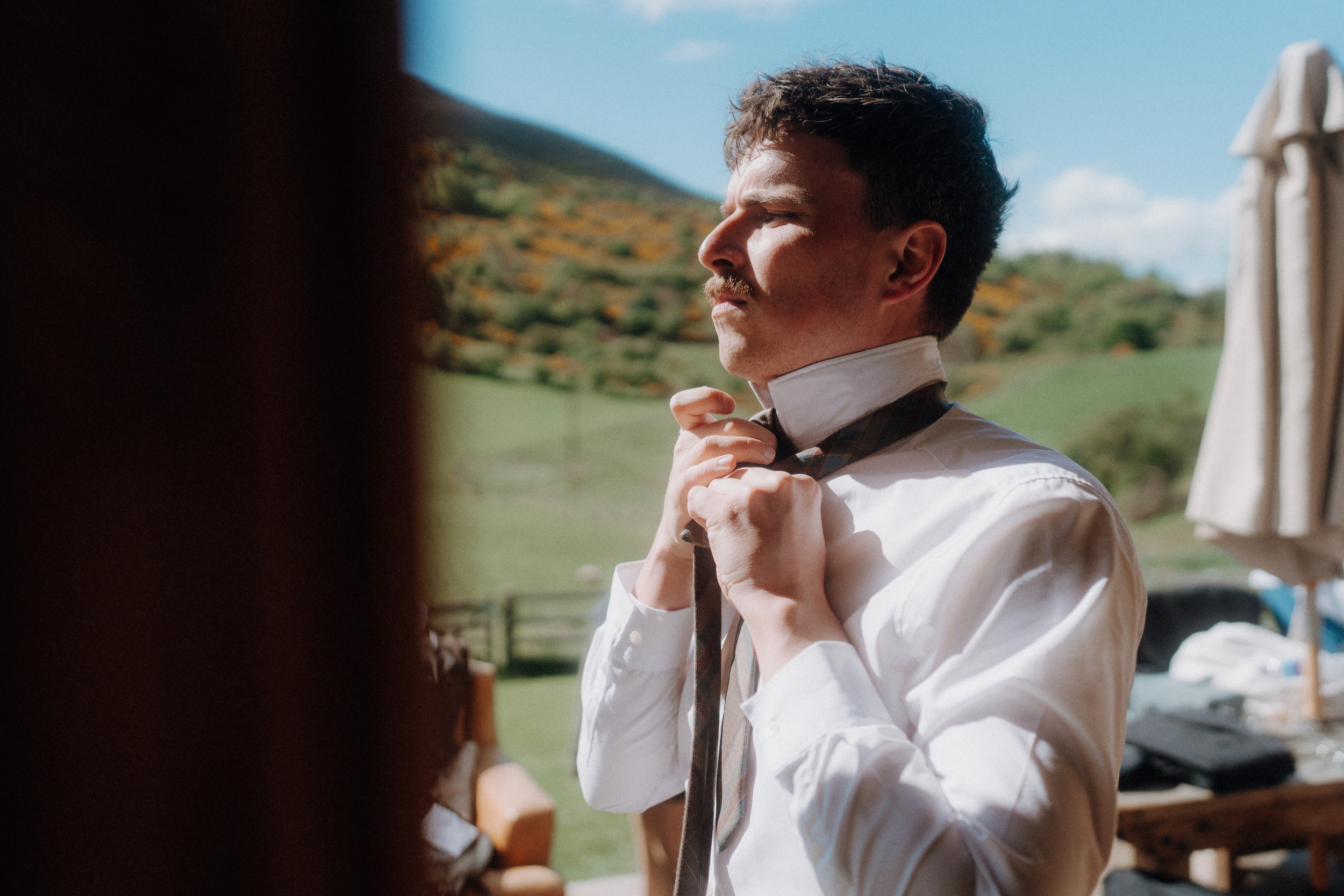 A man in a white shirt adjusts his tie while standing outdoors, with green hills and a blue sky visible in the background.