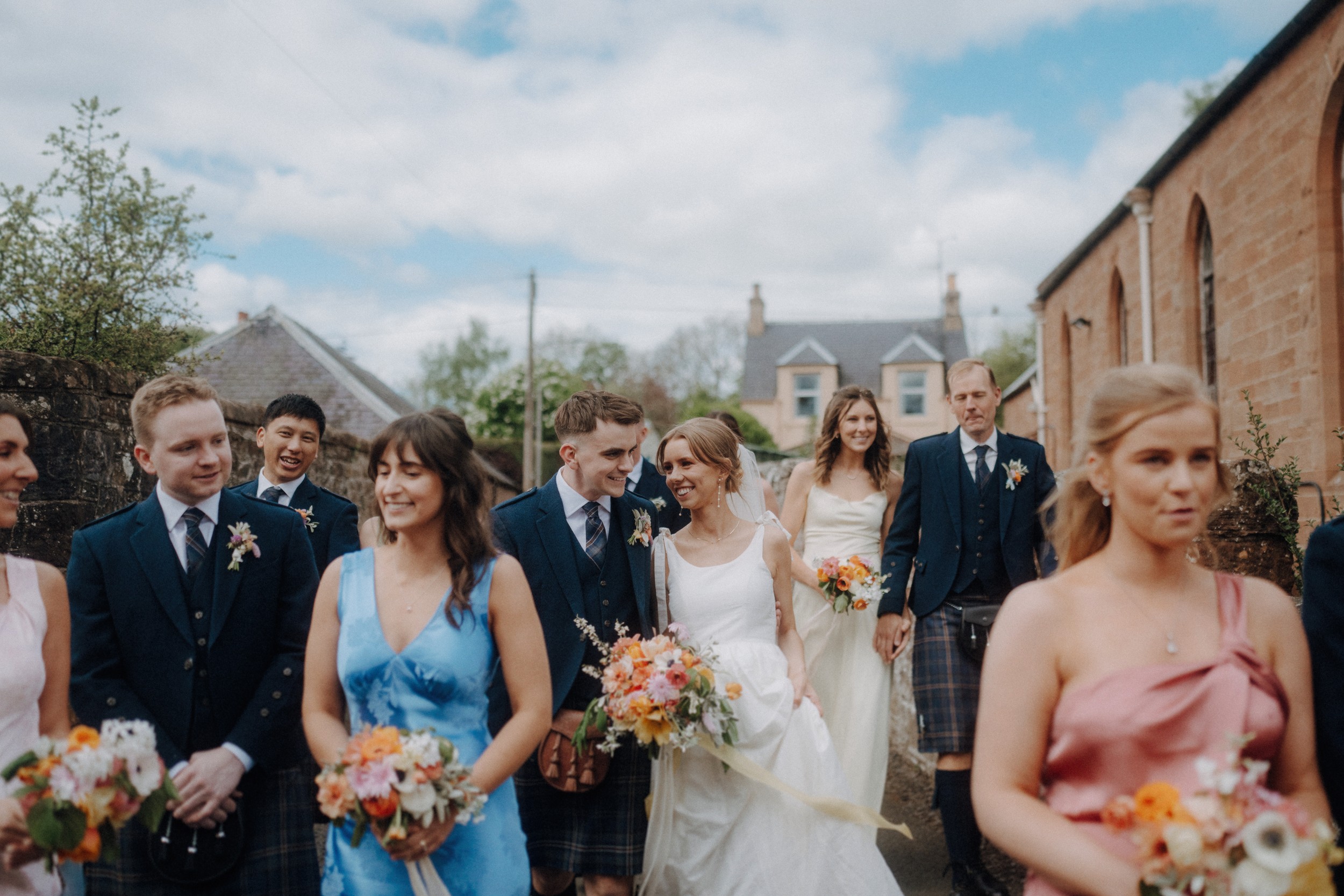 A bride and groom walk outdoors with their wedding party, who are dressed in formal attire with floral bouquets. The group appears to be smiling and enjoying the moment.