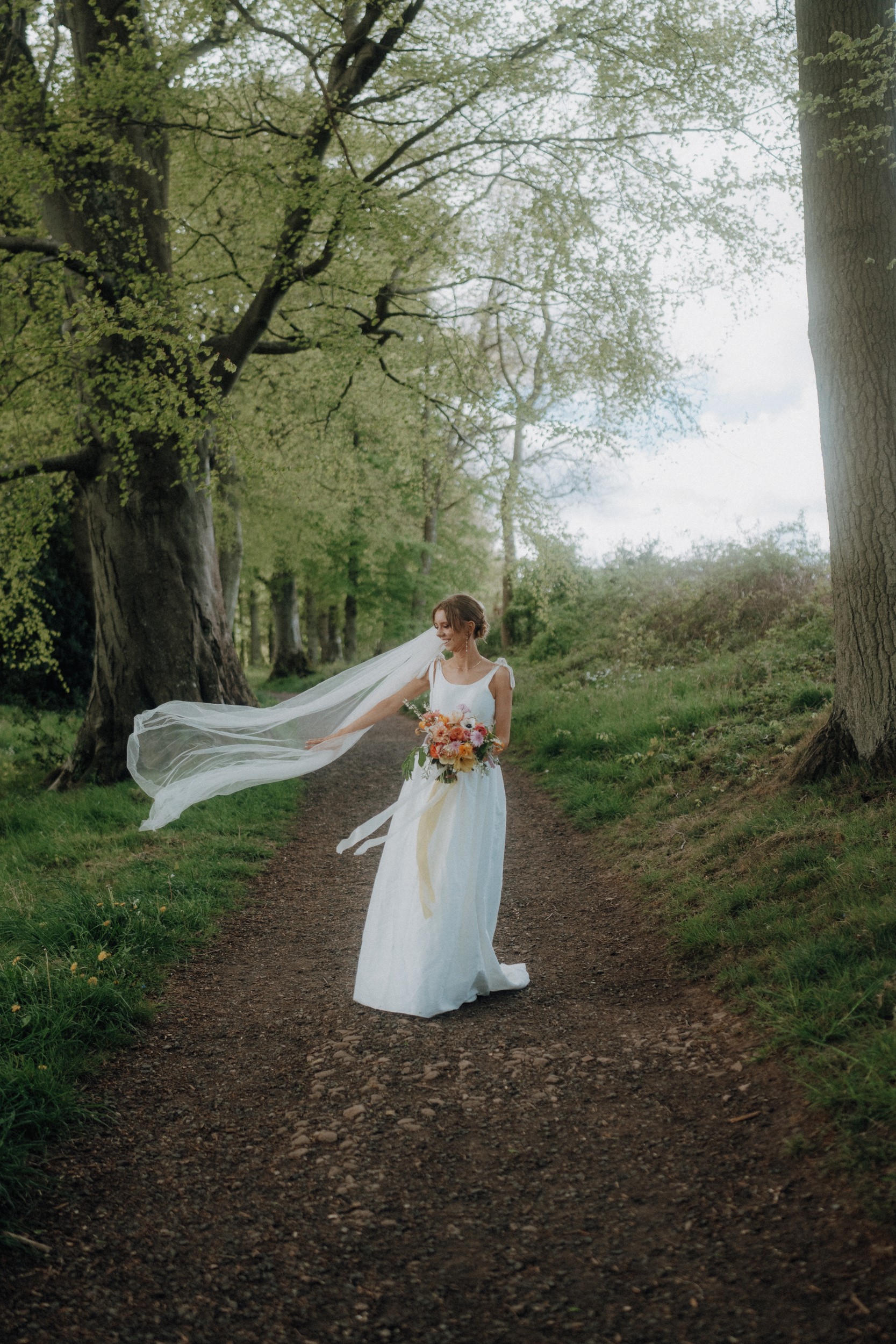A woman in a white wedding dress holds a bouquet while standing on a tree-lined path, her veil blowing in the wind.