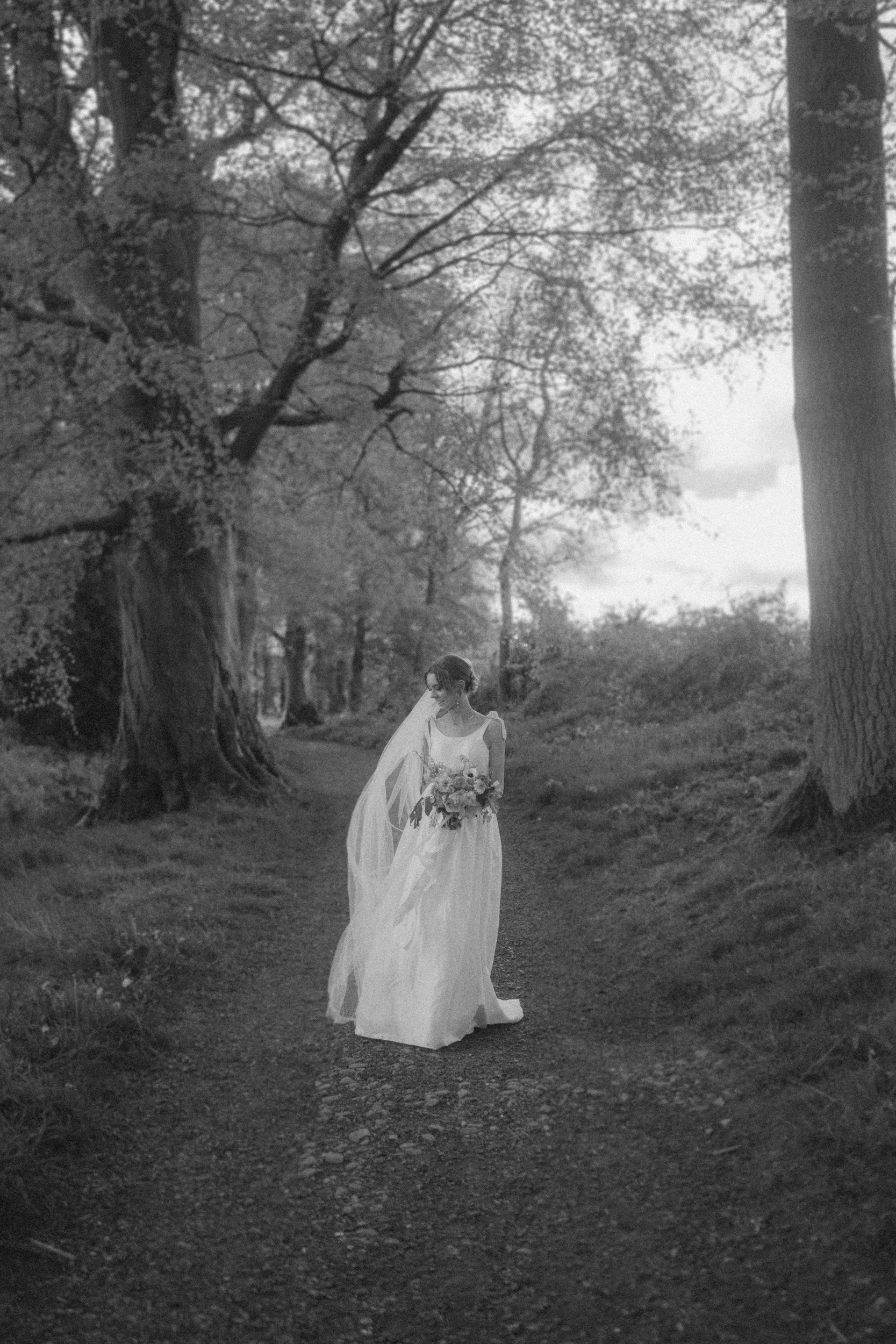A bride in a long white dress and veil holding a bouquet walks alone down a wooded path, surrounded by tall trees. The photo is in black and white.