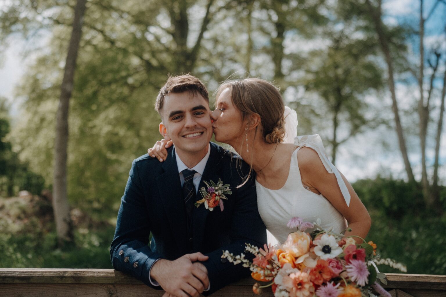 A bride in a white dress kisses the groom on the cheek as he smiles, with a bouquet of flowers and greenery in the background.
