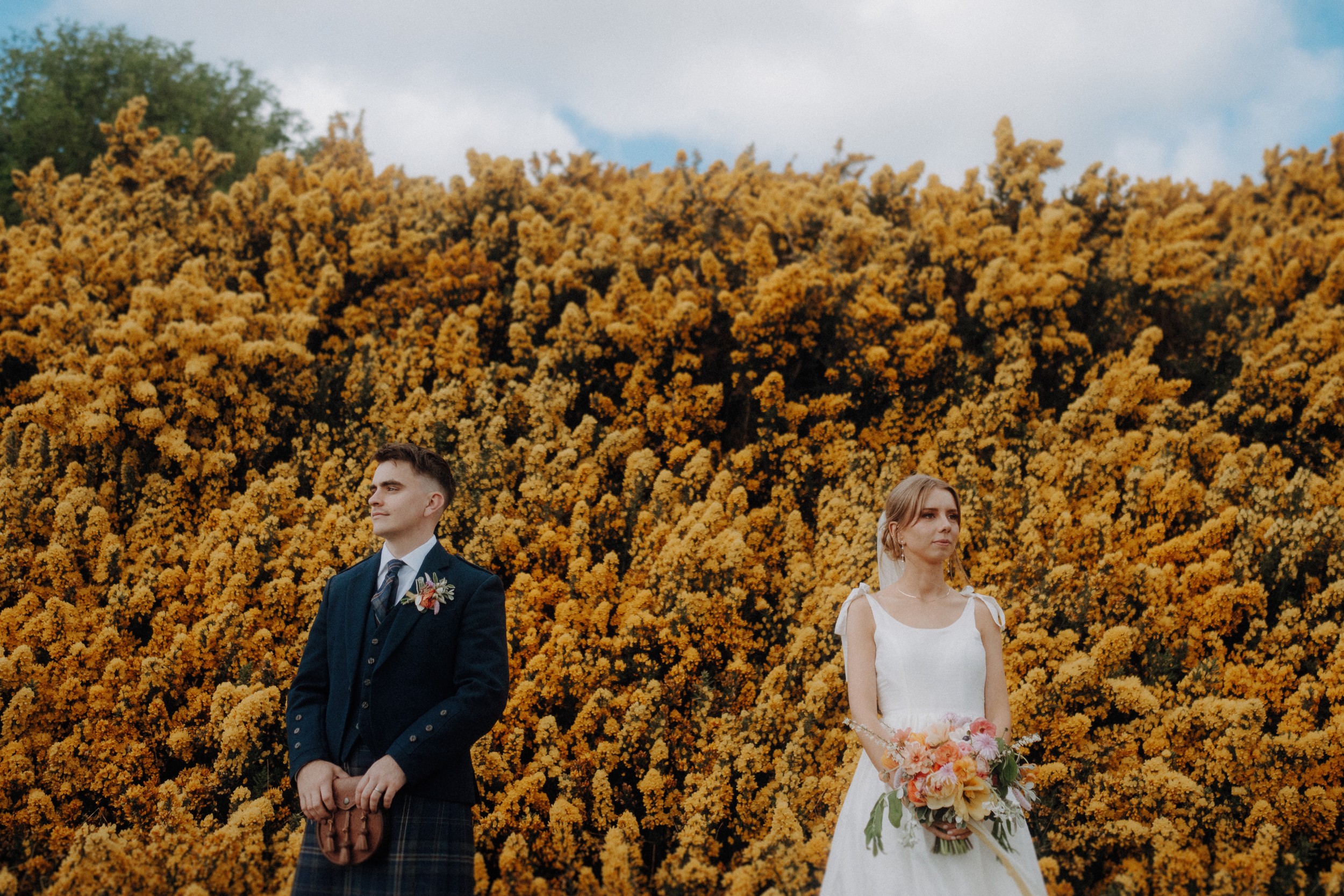 A man and woman in formal attire stand apart in front of dense yellow flowering bushes, both holding floral arrangements and looking in opposite directions.