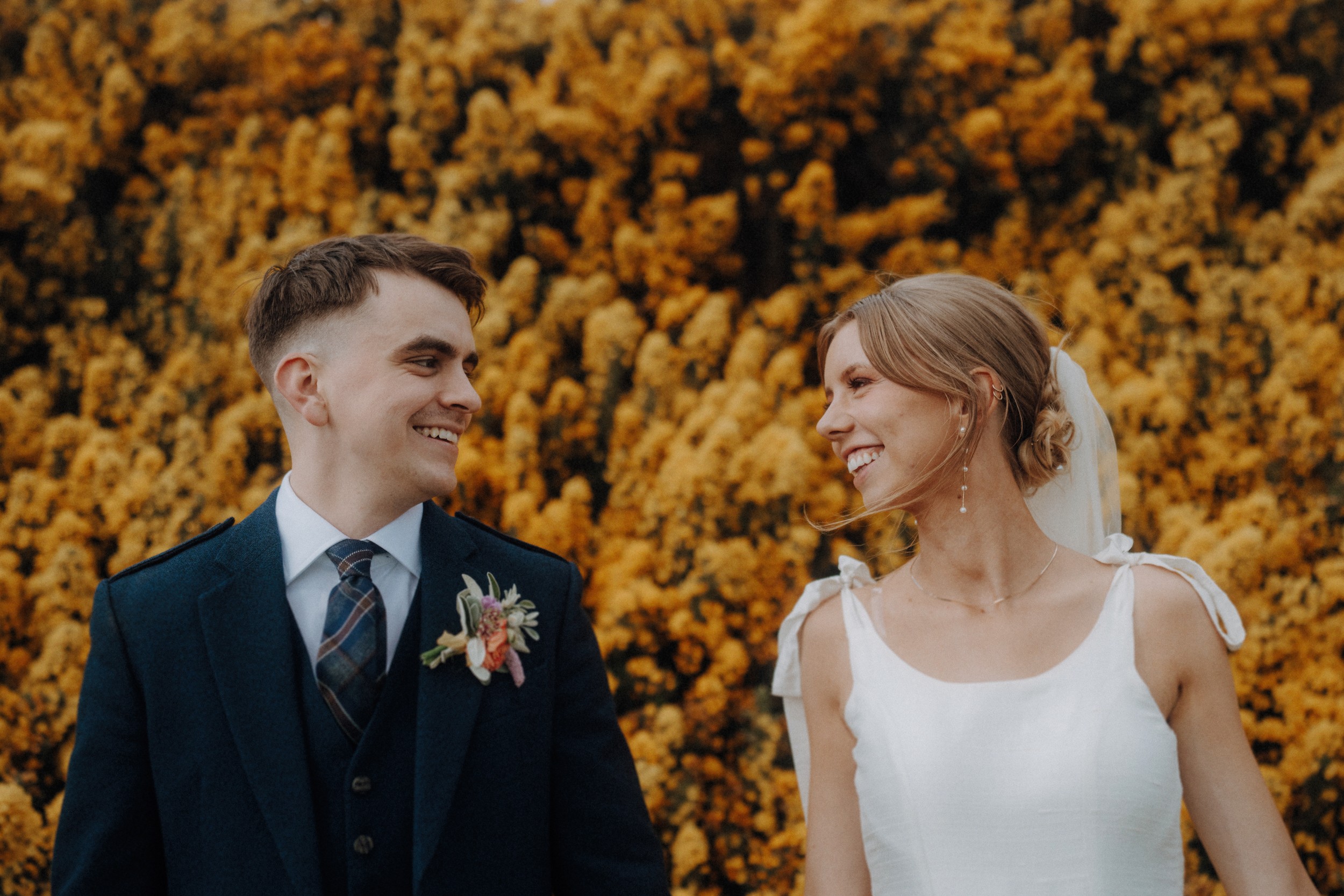 A bride and groom stand outdoors, smiling at each other in front of a backdrop of yellow flowers. The groom wears a dark suit; the bride wears a white dress and veil.