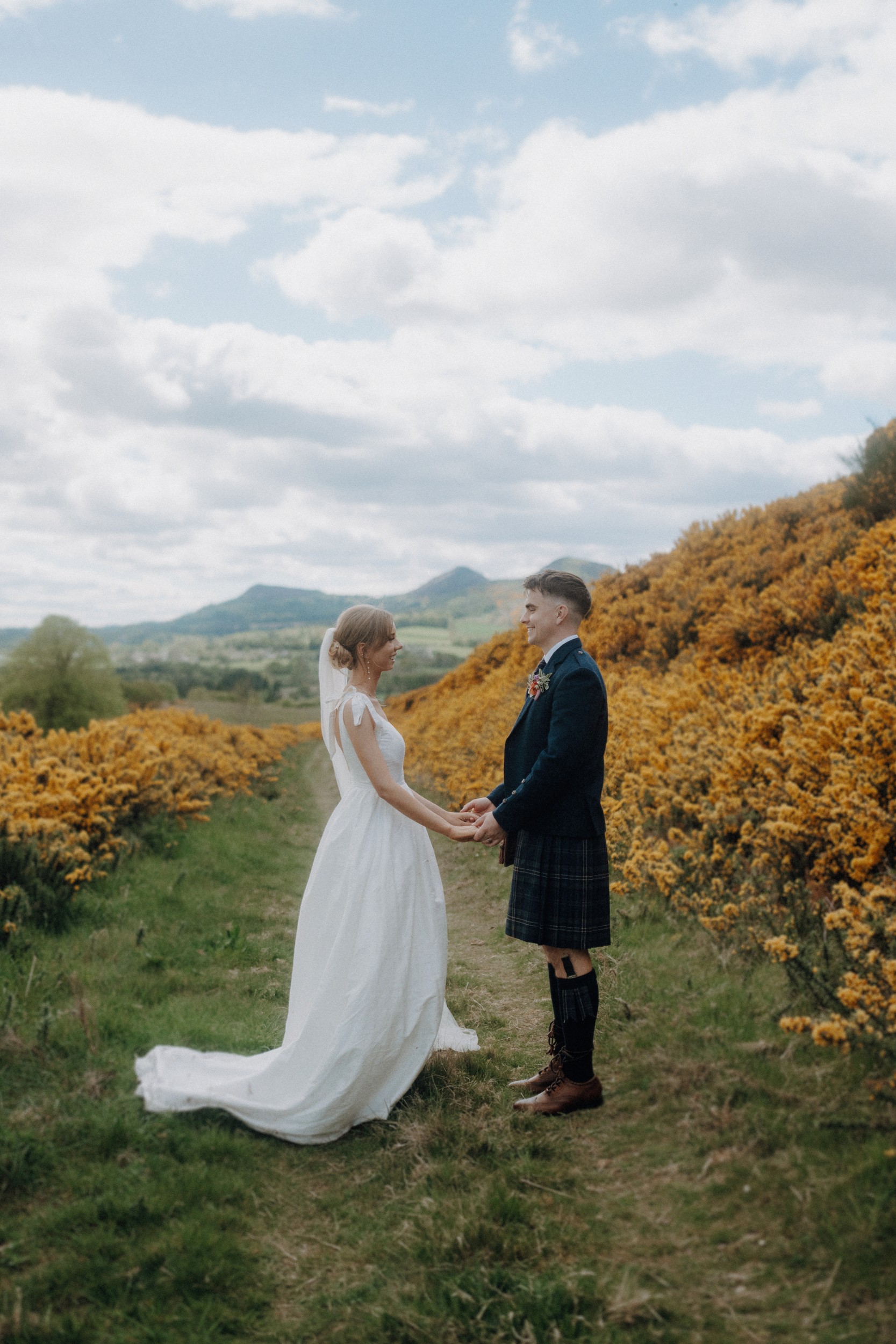 A bride and groom stand facing each other, holding hands on a grassy path bordered by yellow flowers, with hills and a cloudy sky in the background.