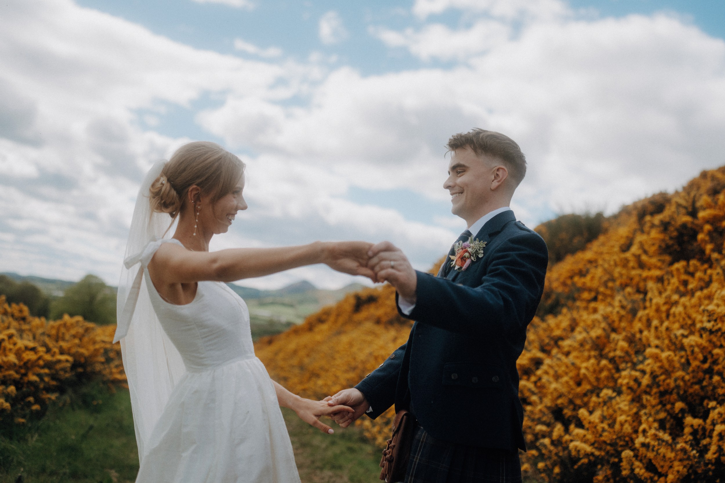 A bride and groom hold hands and smile at each other outdoors, surrounded by yellow flowering bushes under a partly cloudy sky.