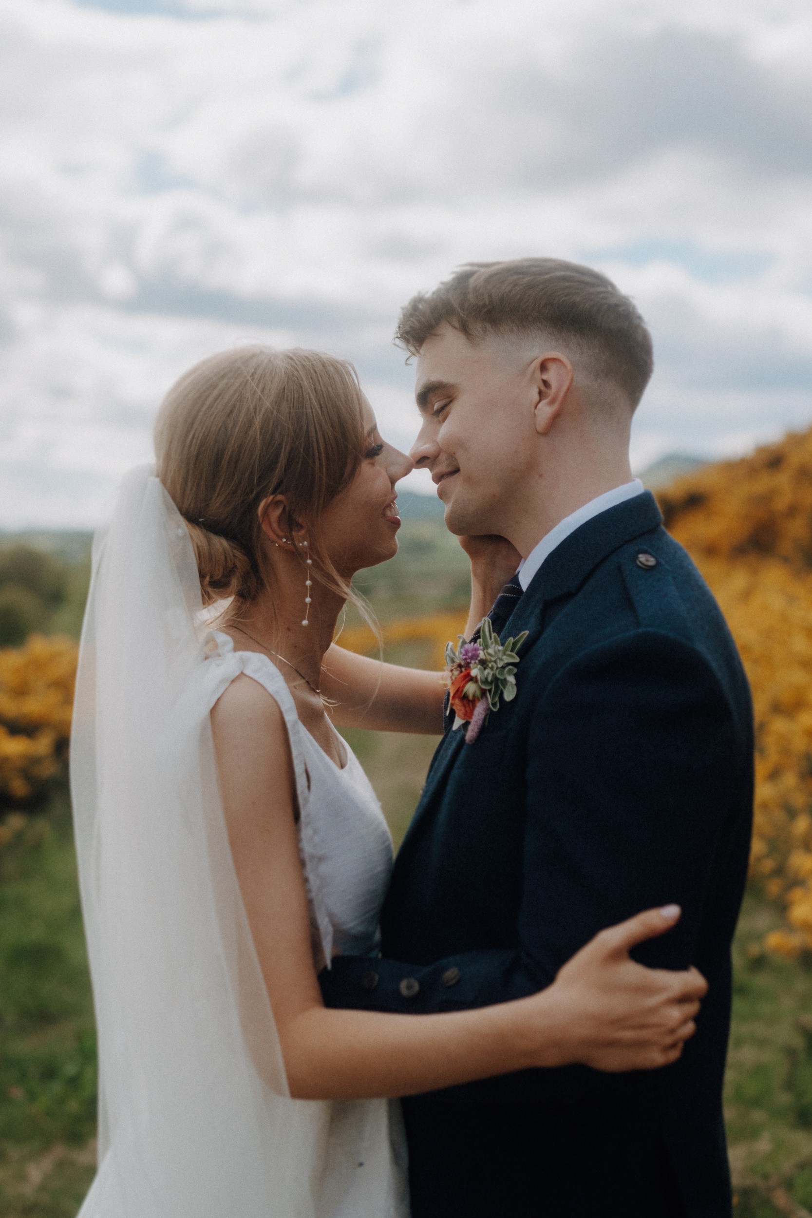 A bride and groom stand outdoors, facing each other and smiling, with the bride in a white dress and veil and the groom in a dark suit, surrounded by greenery and yellow flowers.