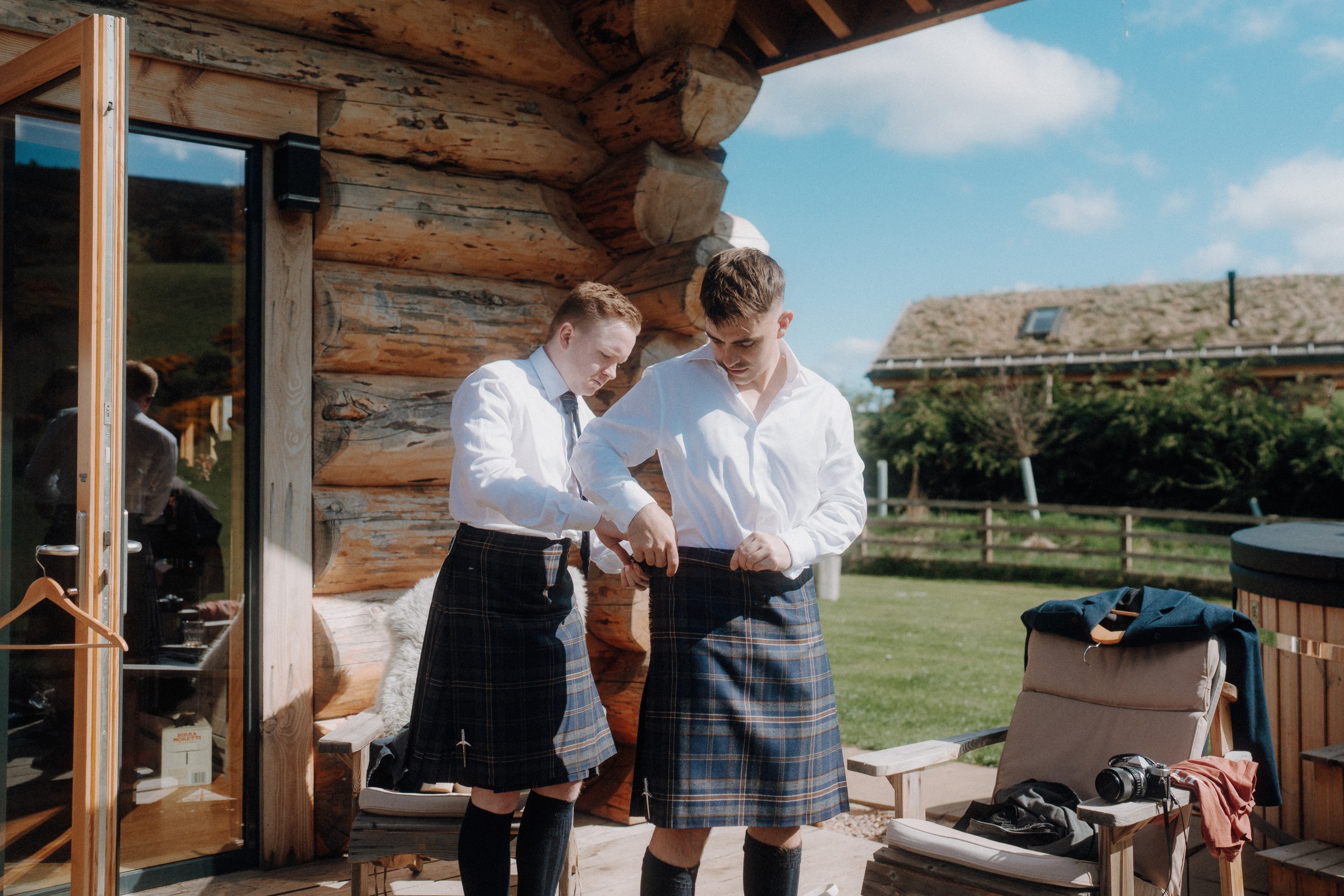Two men in white shirts and tartan kilts stand outside a wooden cabin, adjusting their outfits on a sunny day.