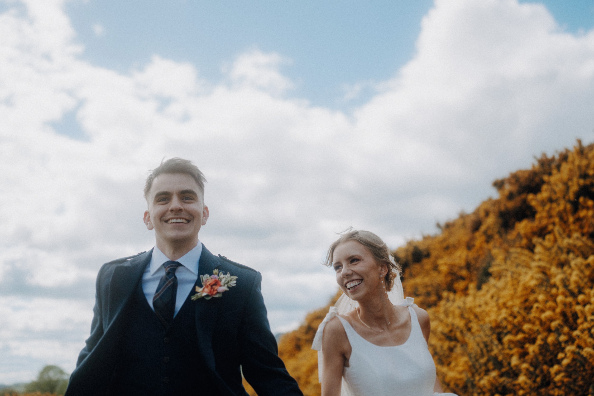 A man in a dark suit and a woman in a white dress smile and walk outdoors, with yellow foliage and a cloudy sky in the background.