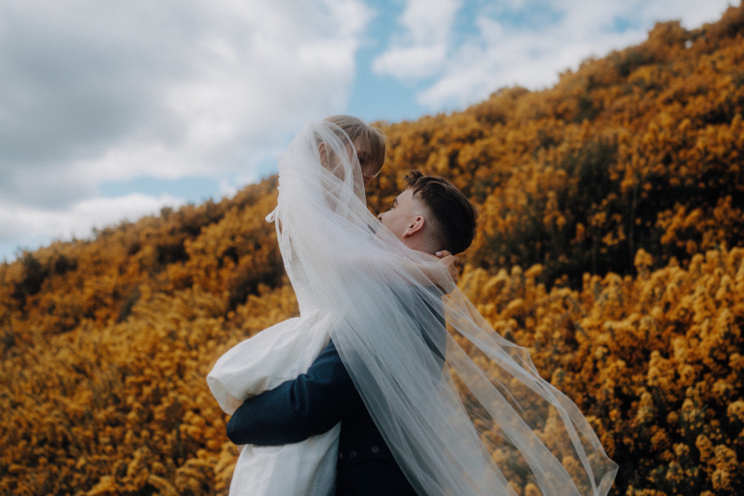 A groom lifts a bride in a white dress and veil outdoors, with a hillside covered in yellow flowers and a cloudy sky in the background.
