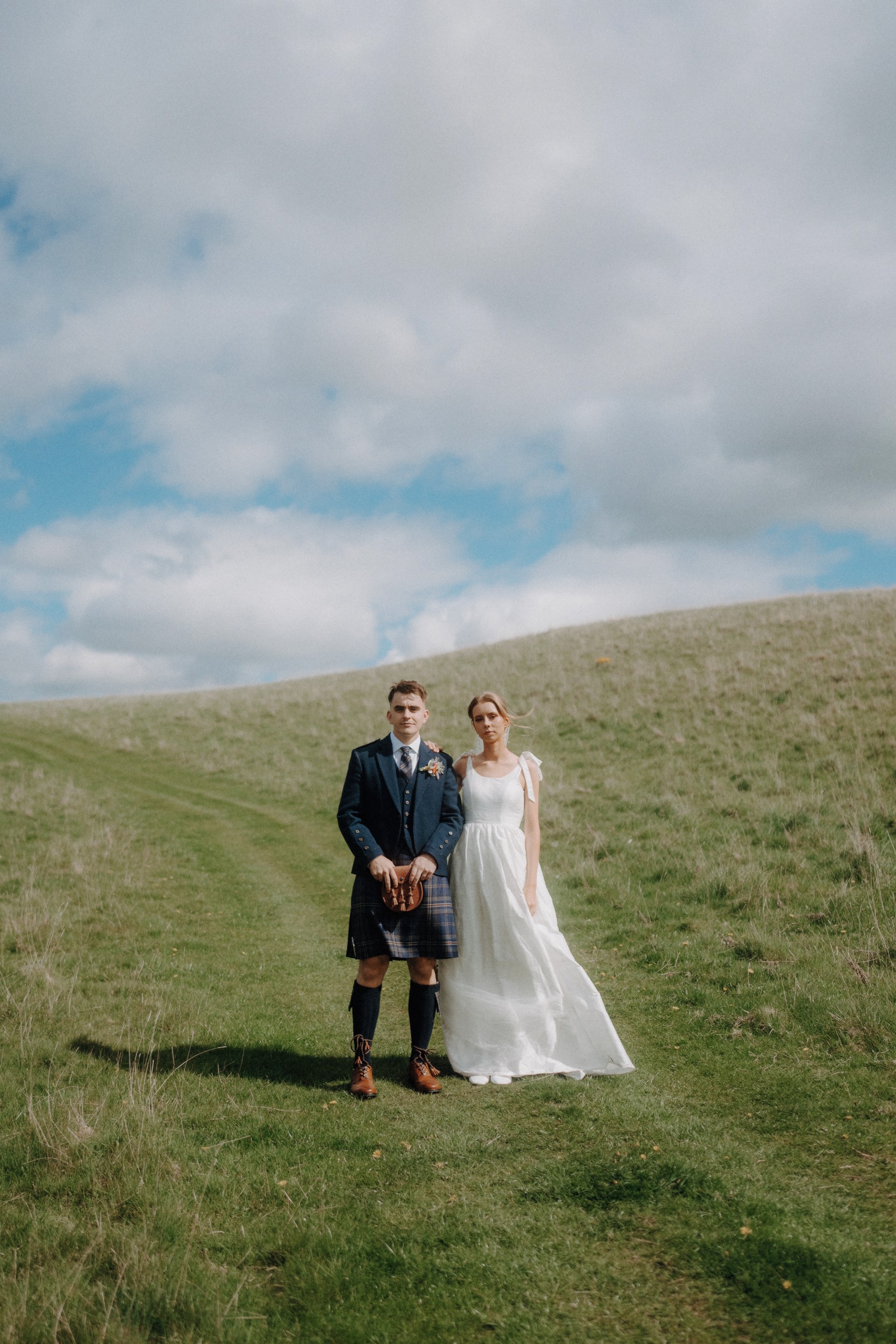 A bride in a white dress and a groom in a kilt stand side by side on a grassy hill under a partly cloudy sky.