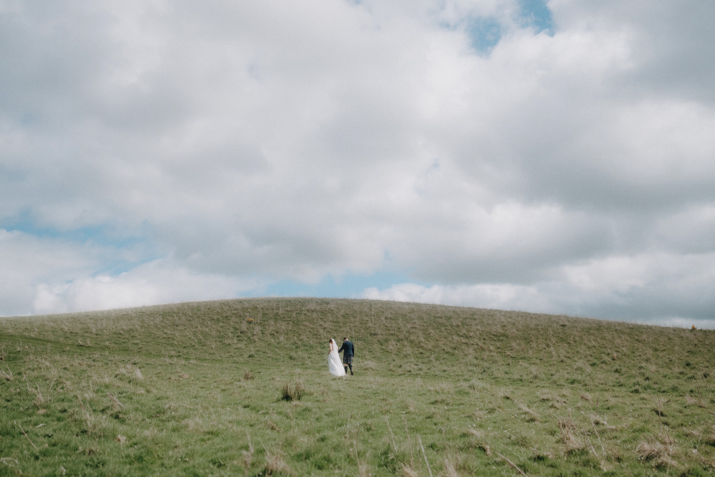 A couple walks hand in hand up a grassy hill under a cloudy sky.