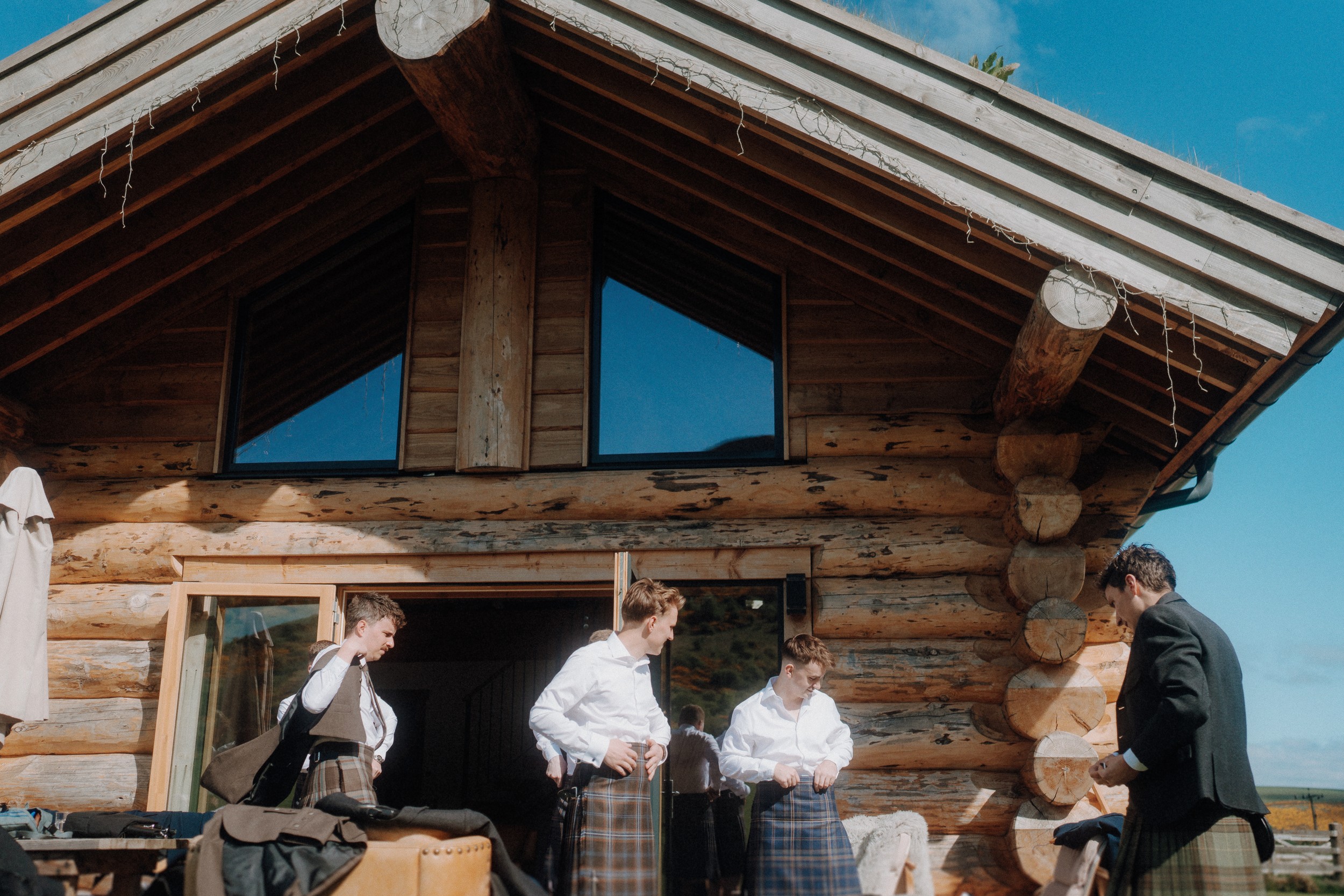 Four men wearing kilts and white shirts stand outside a wooden log cabin, adjusting their outfits under a bright blue sky.