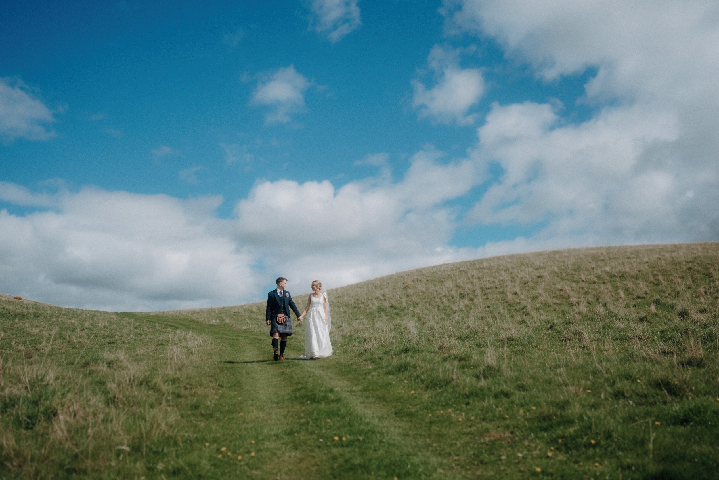 A couple in formal attire walks hand in hand along a grassy path between rolling hills under a partly cloudy blue sky.