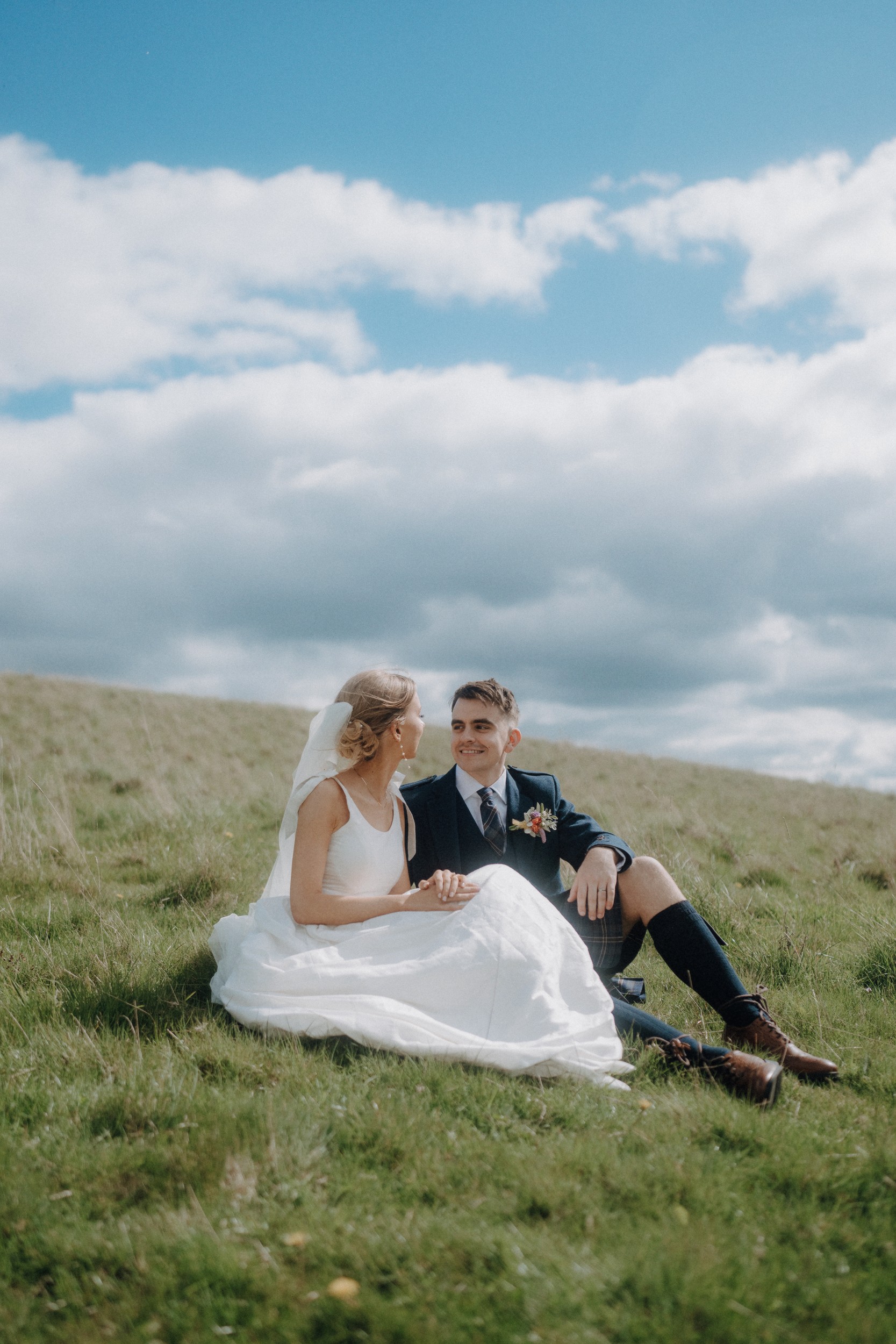 A bride and groom sit together on a grassy hillside under a partly cloudy sky, dressed in wedding attire, looking at each other and smiling.
