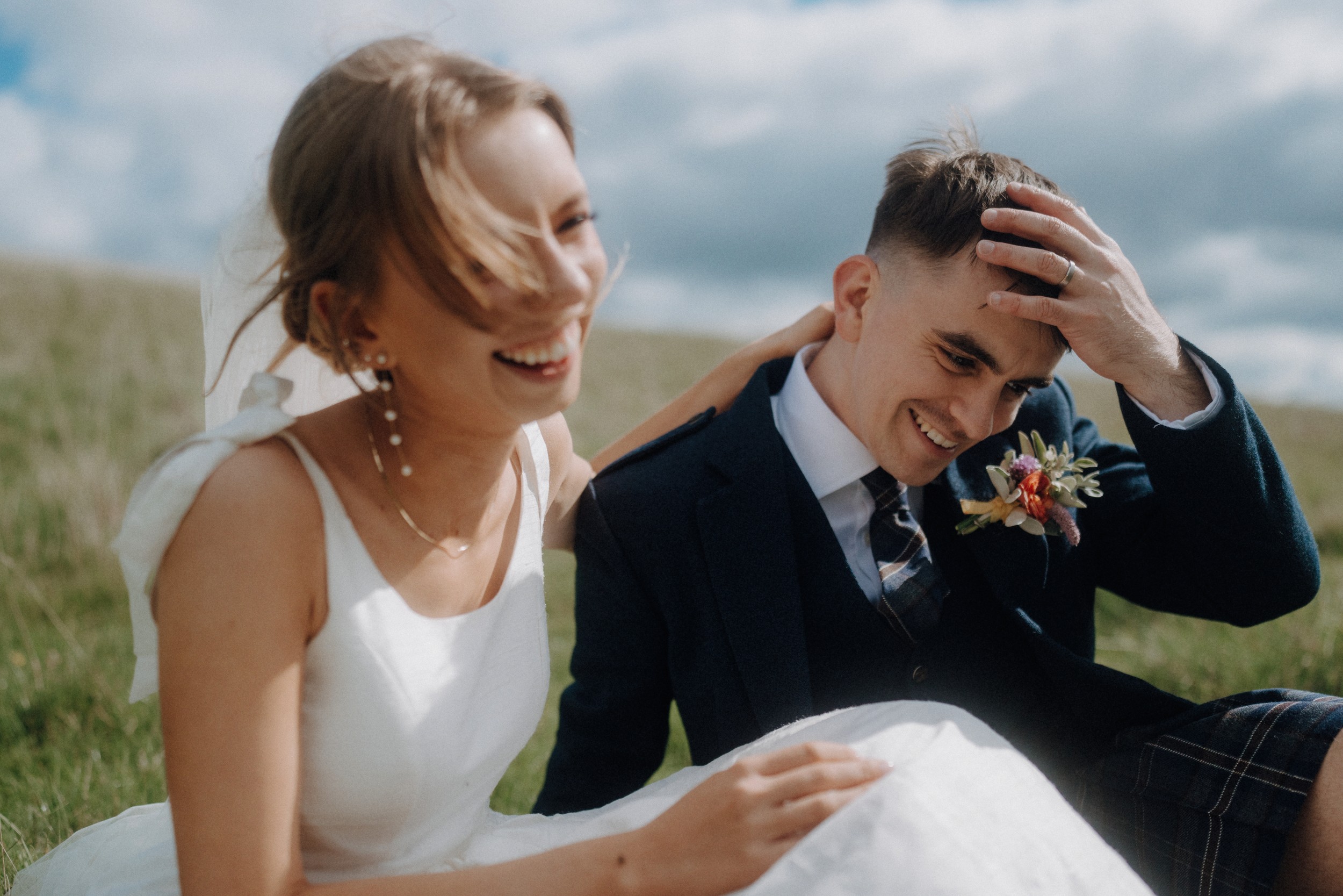 A bride and groom sit on grass outdoors, smiling and laughing together. The bride wears a white dress; the groom wears a suit with a boutonnière.