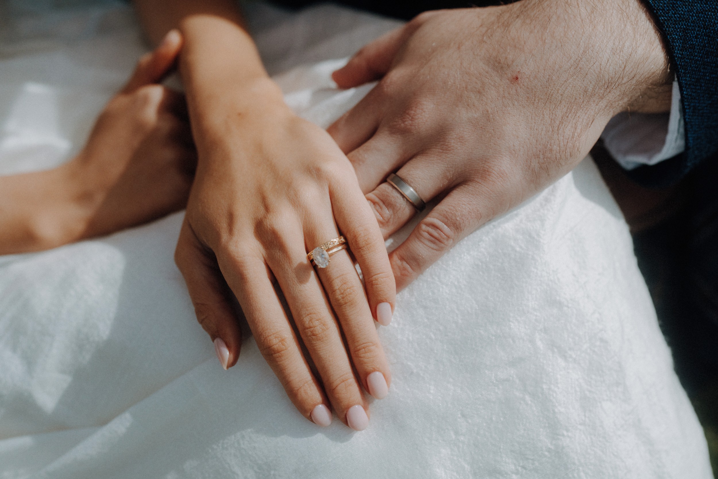 Close-up of two hands resting together, one wearing a gold ring with a diamond and the other wearing a plain silver band, both on a white fabric background.