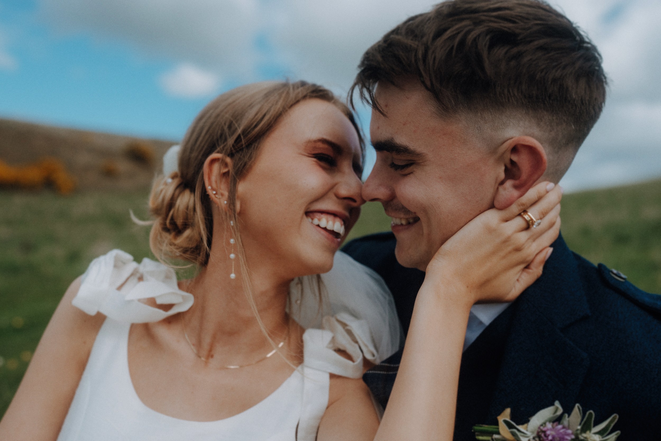 A bride and groom smile and touch foreheads outdoors, with greenery and a cloudy sky in the background.
