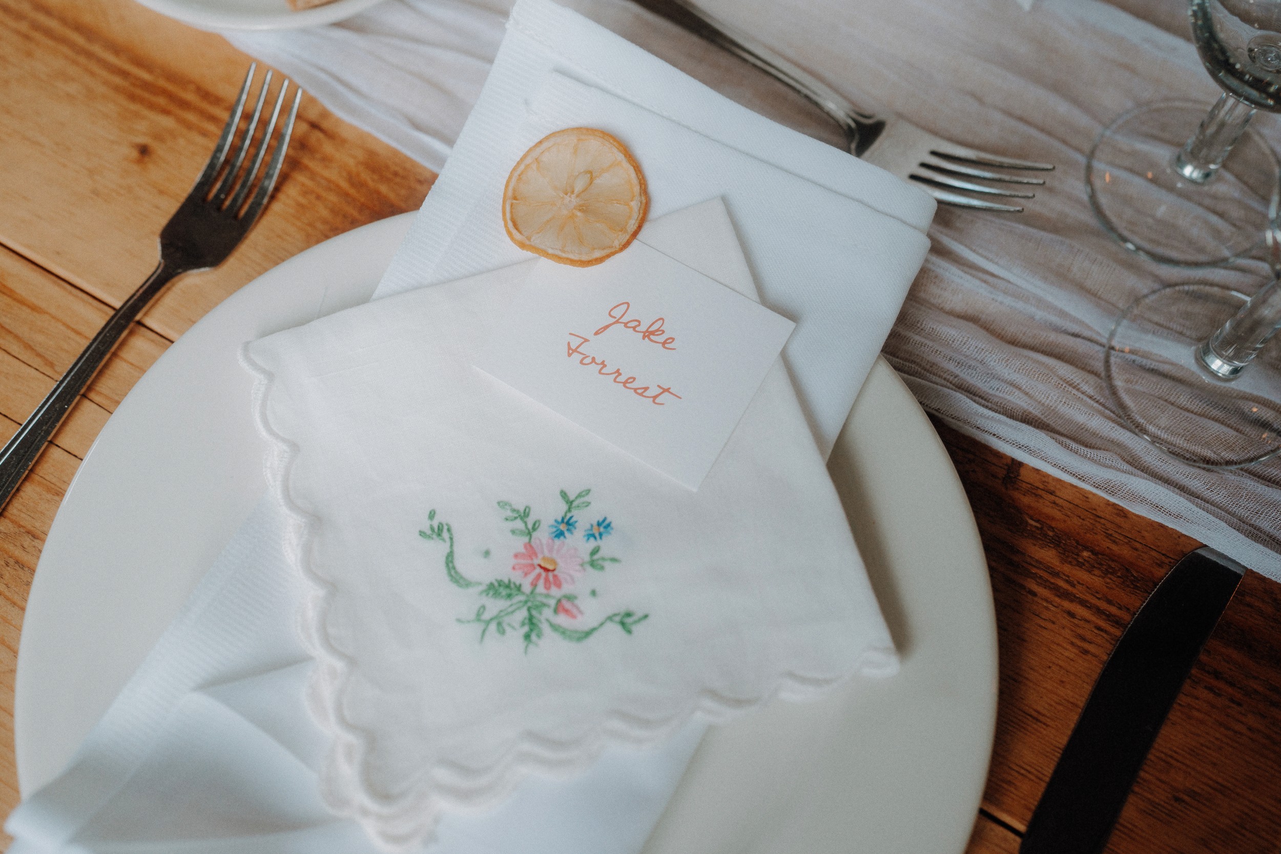 Place setting with a white plate, embroidered napkin, name card, dried lemon slice, fork, knife, and glasses on a wooden table.