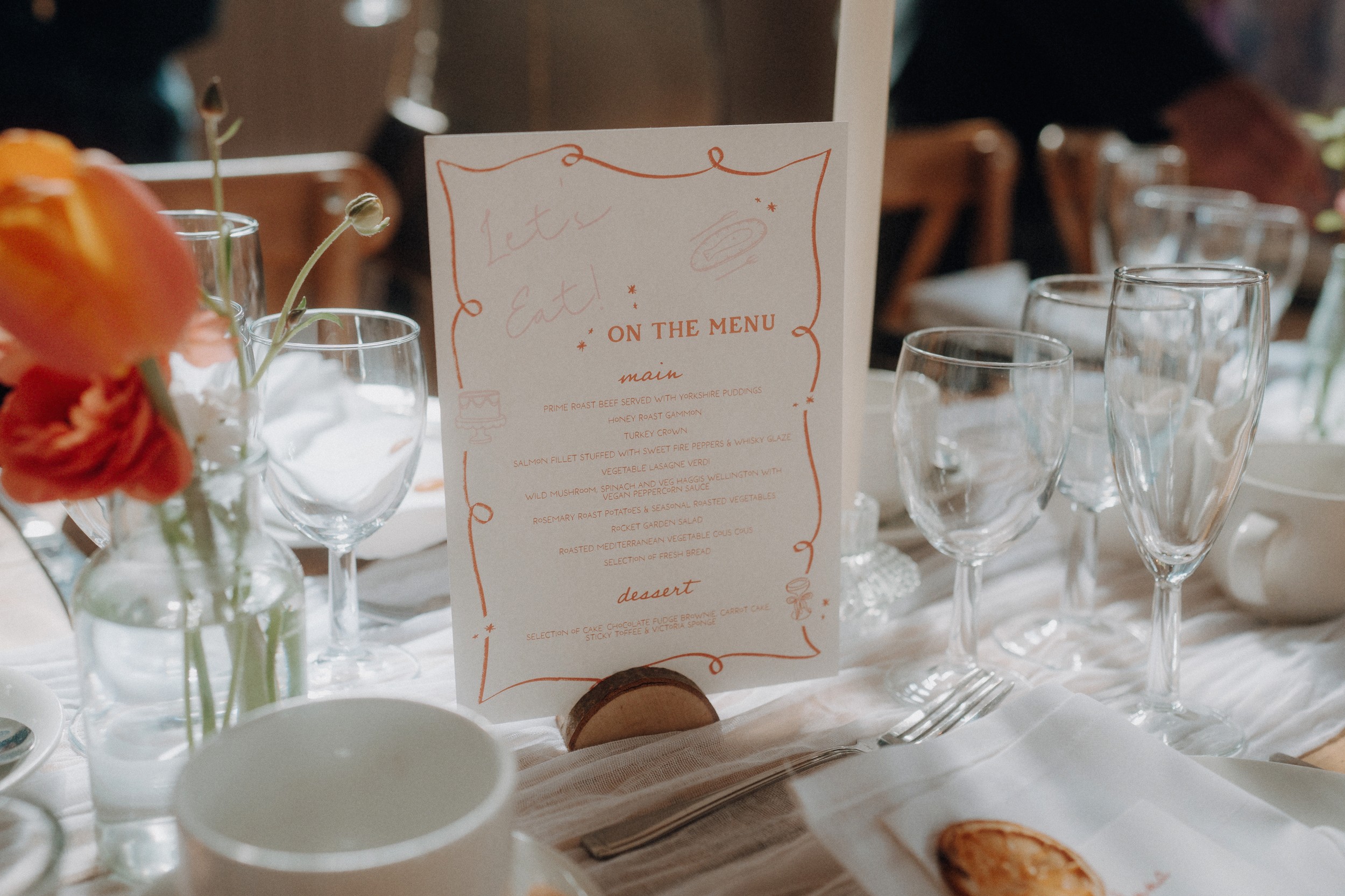 A wedding table setting with glassware, cutlery, a flower vase, and a printed menu card displaying the food options.