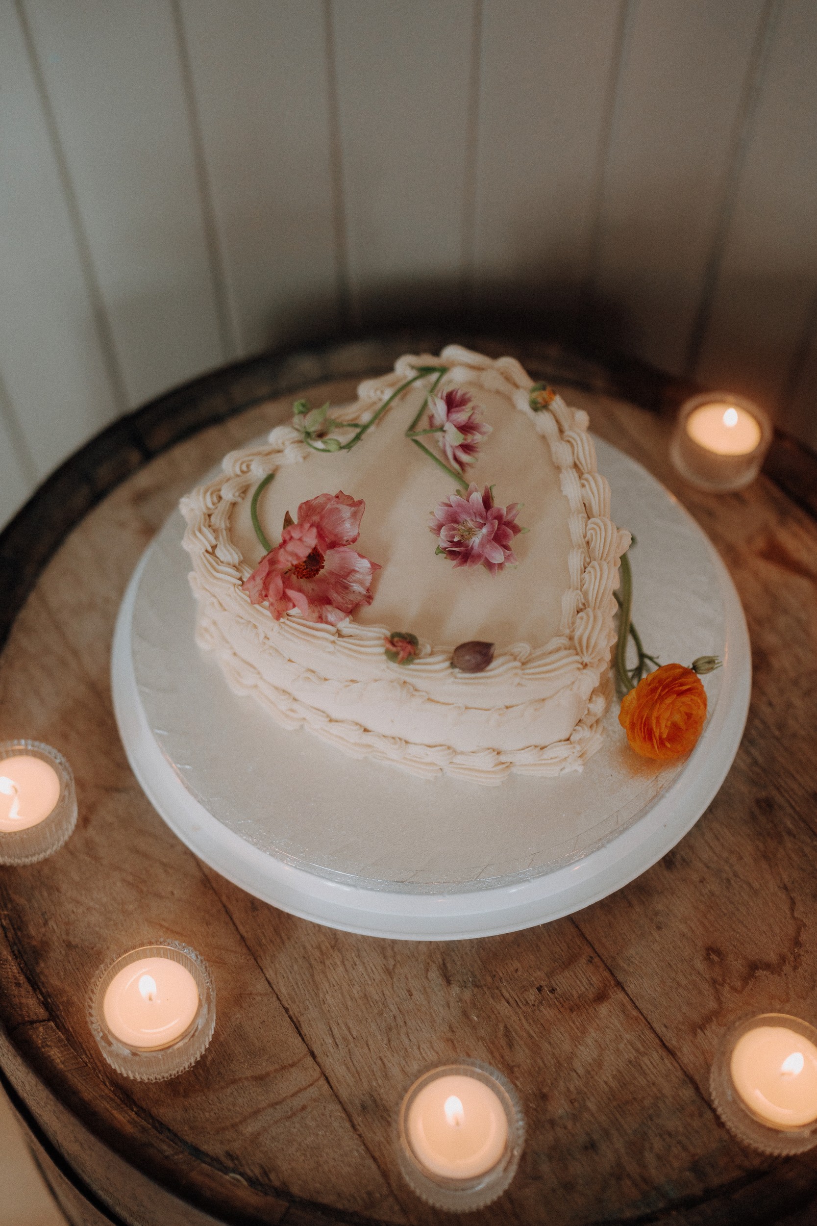 A heart-shaped cake decorated with pink and orange flowers sits on a round wooden table surrounded by small lit tealight candles.