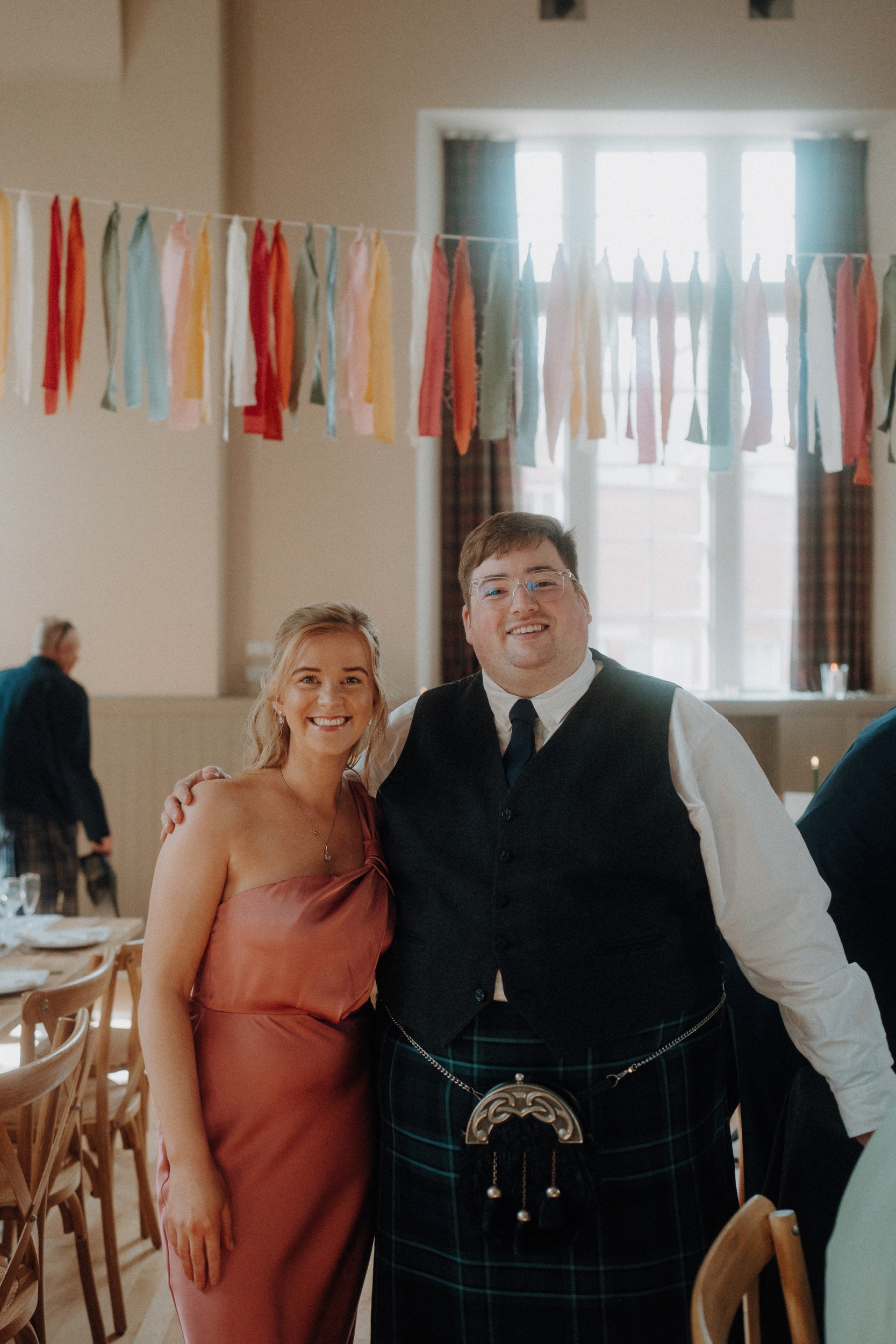 A woman in a pink dress and a man in a kilt and vest stand together smiling in a decorated room with hanging colorful ribbons.