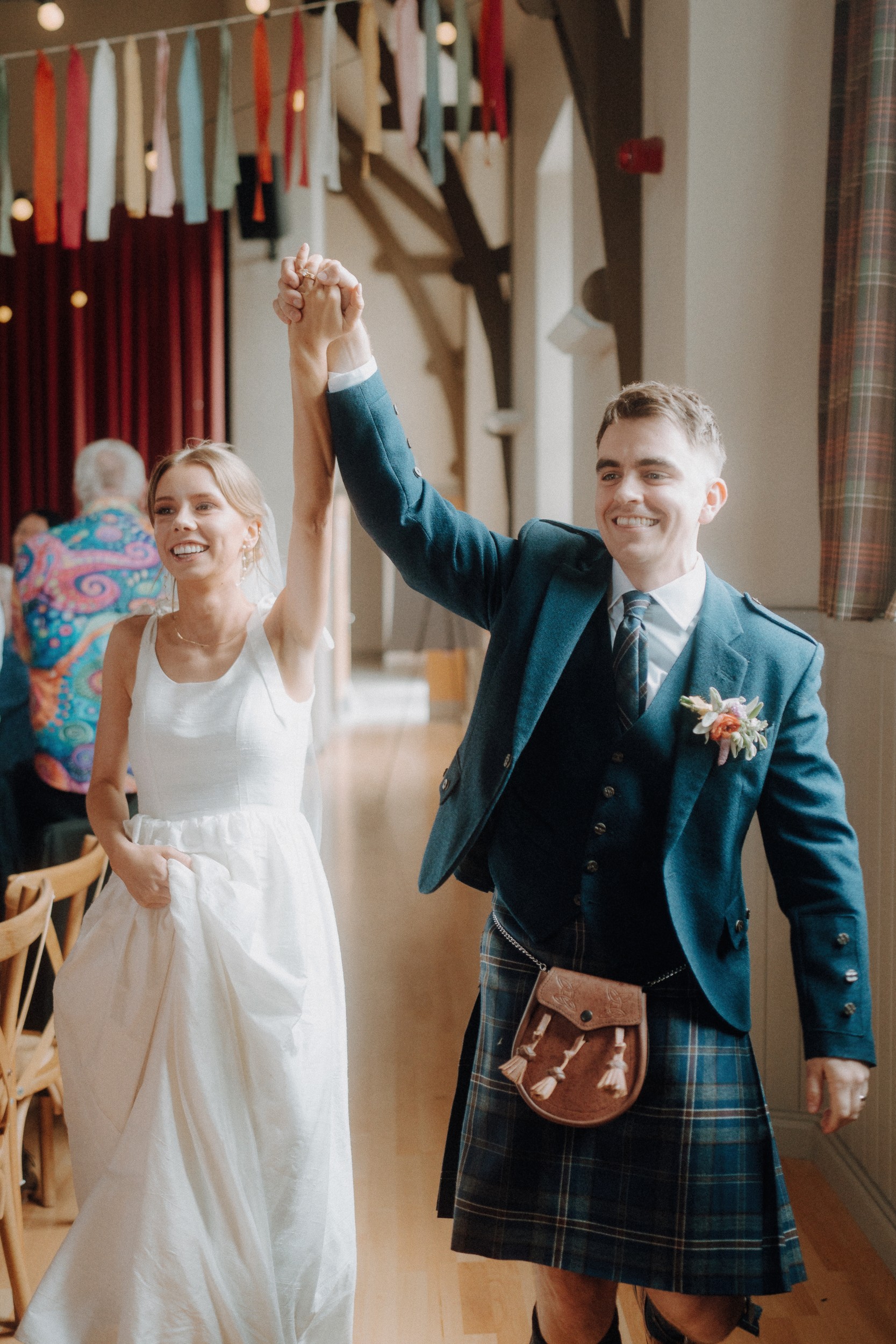 A bride in a white dress and a groom in a kilt walk hand in hand, smiling, at an indoor wedding venue decorated with colorful hanging ribbons.