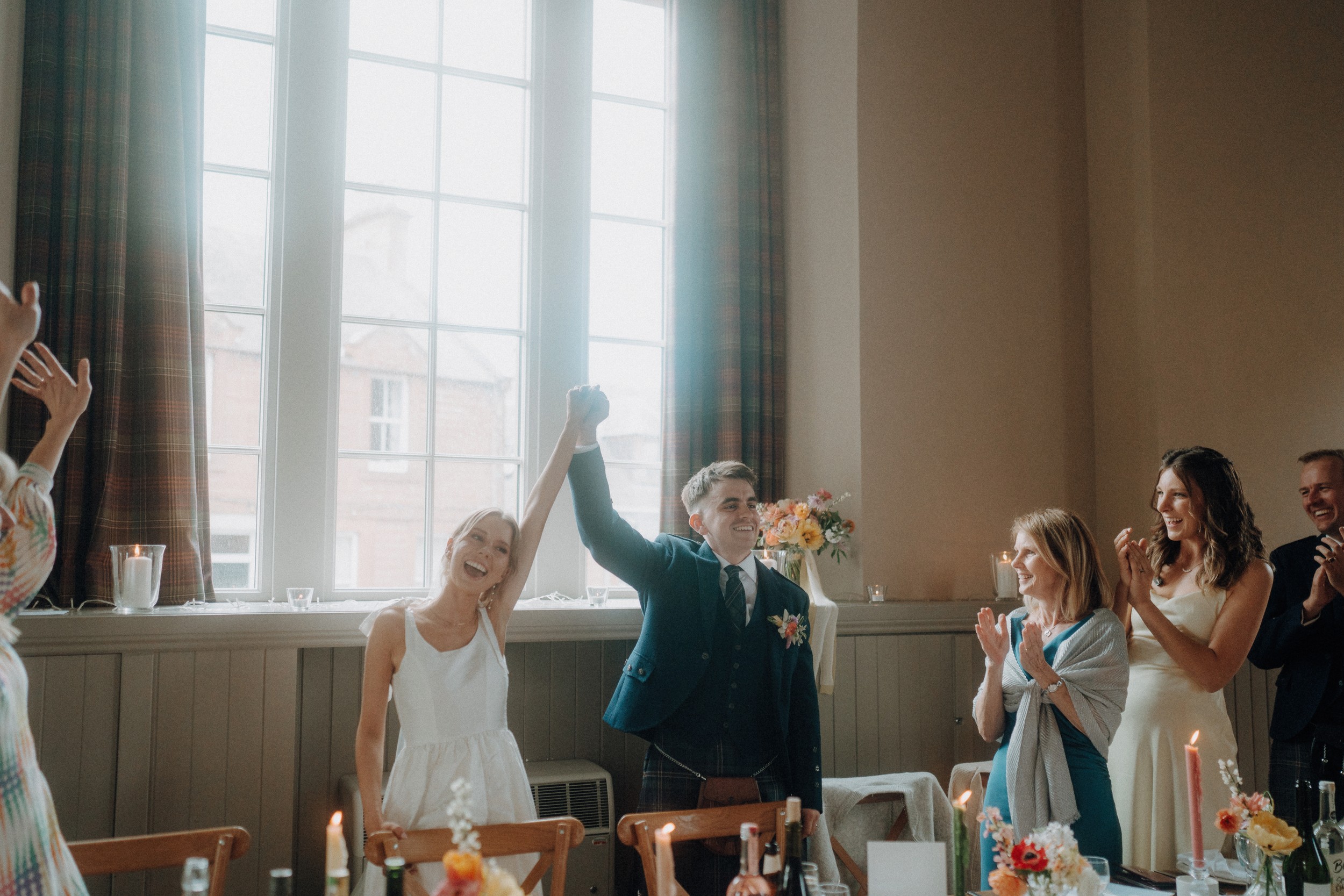 A newlywed couple stands by a table, holding hands raised in celebration, while guests around them applaud in a sunlit room.