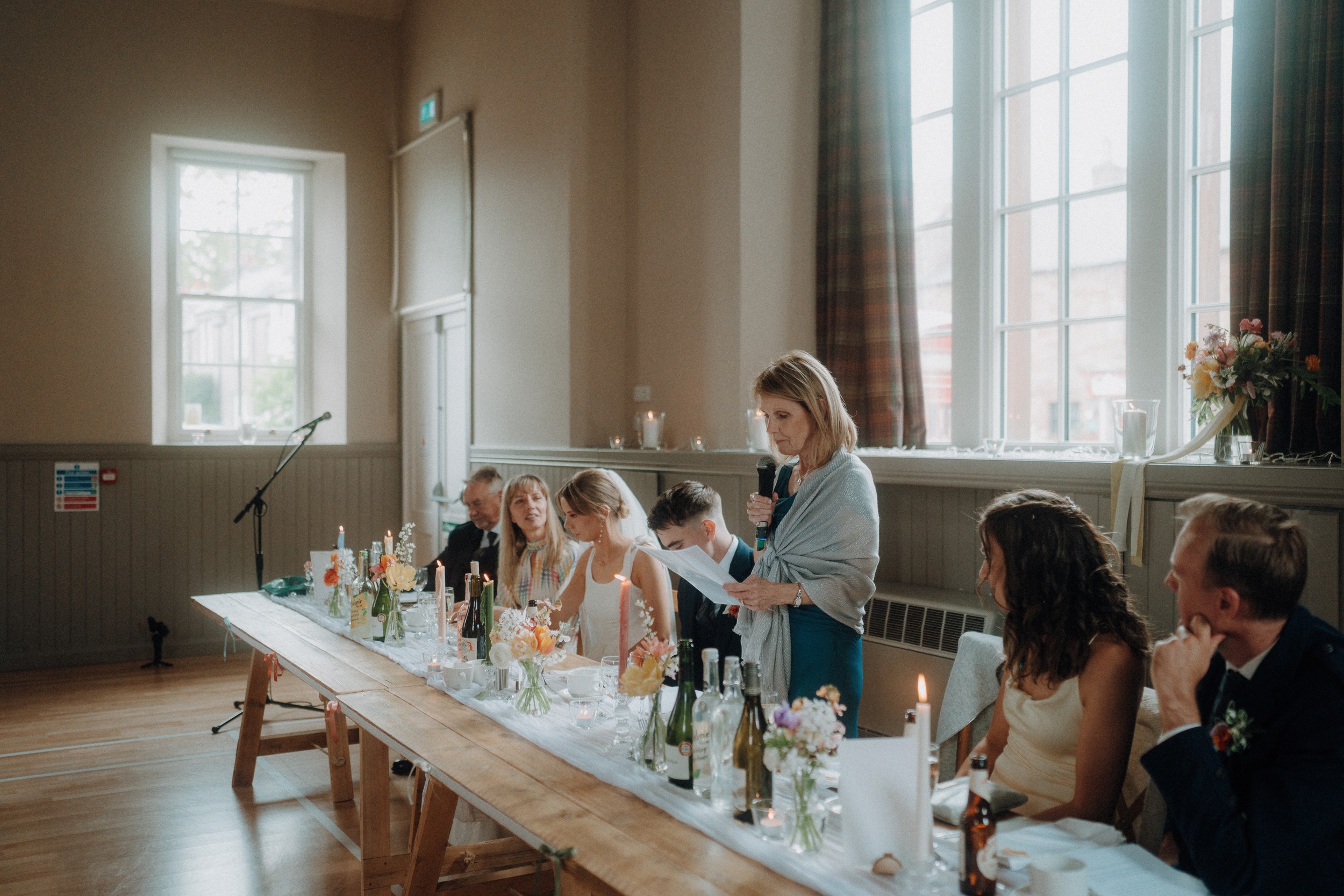 A woman stands and speaks into a microphone at a long table set for a formal event, with several guests seated and listening in a bright, decorated room.