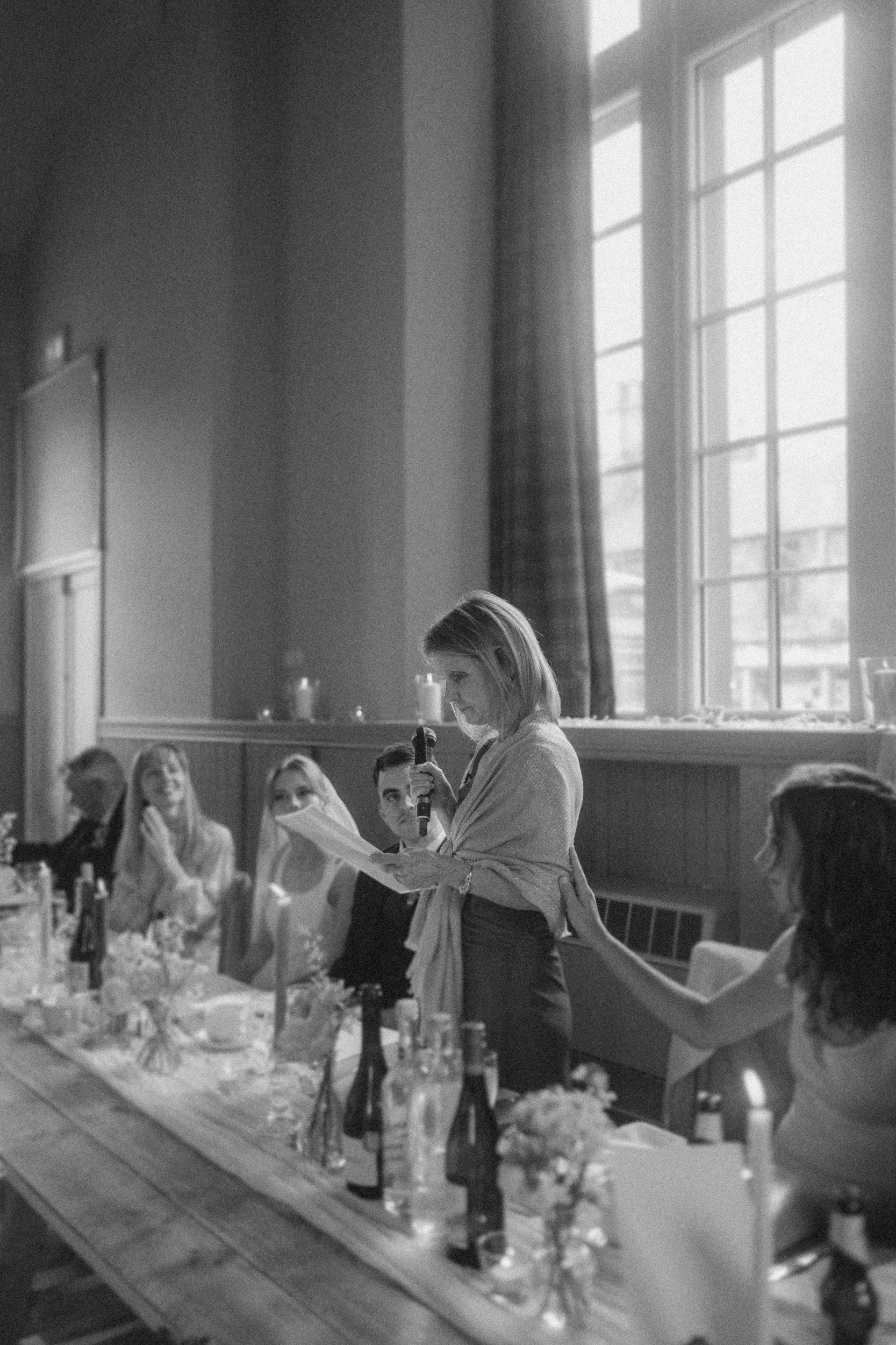 A woman stands and reads from a paper into a microphone at a long table during an indoor gathering, while others are seated and listening.
