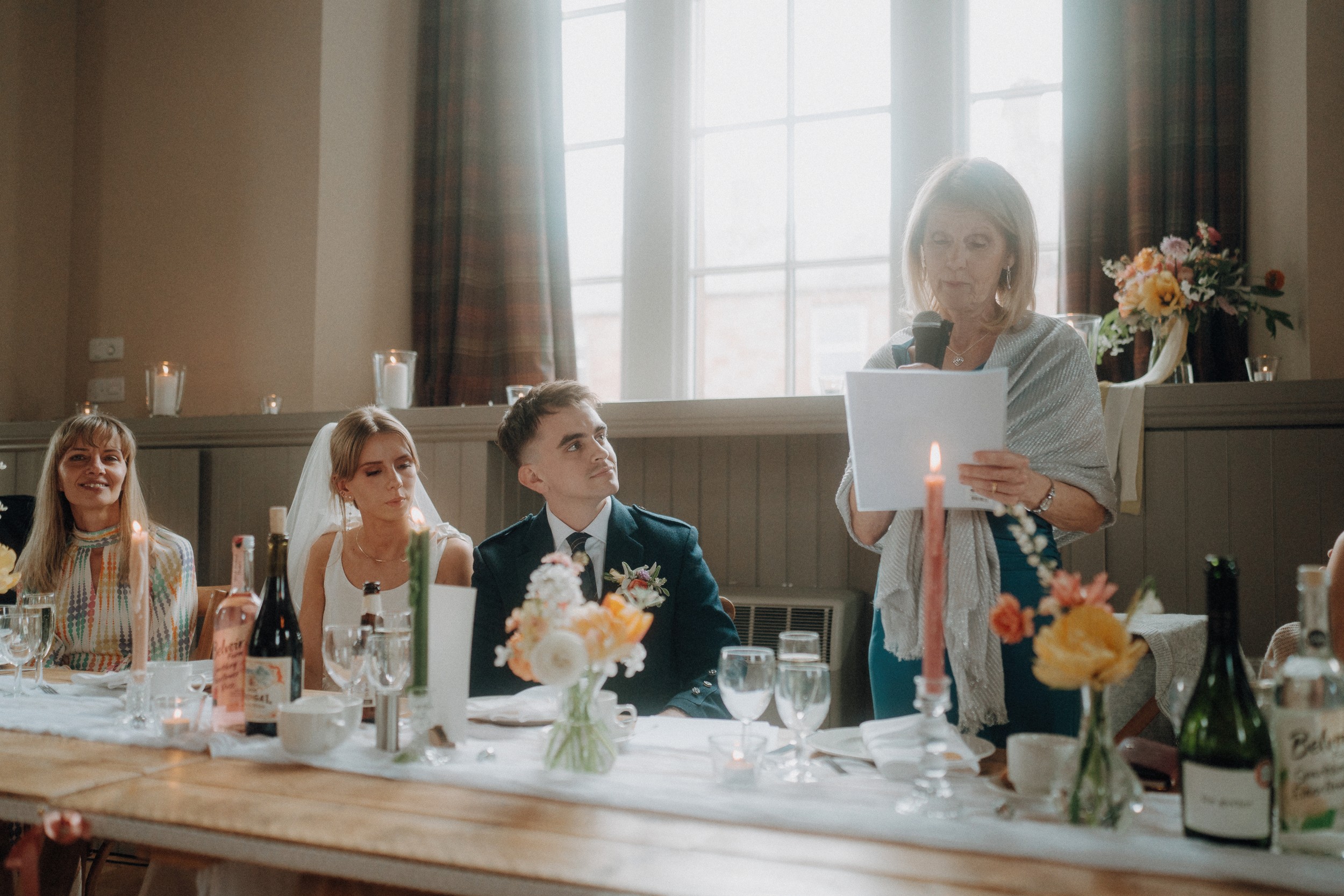 A woman stands and reads from a paper at a table during a wedding reception, with the bride and groom seated beside her and guests listening.