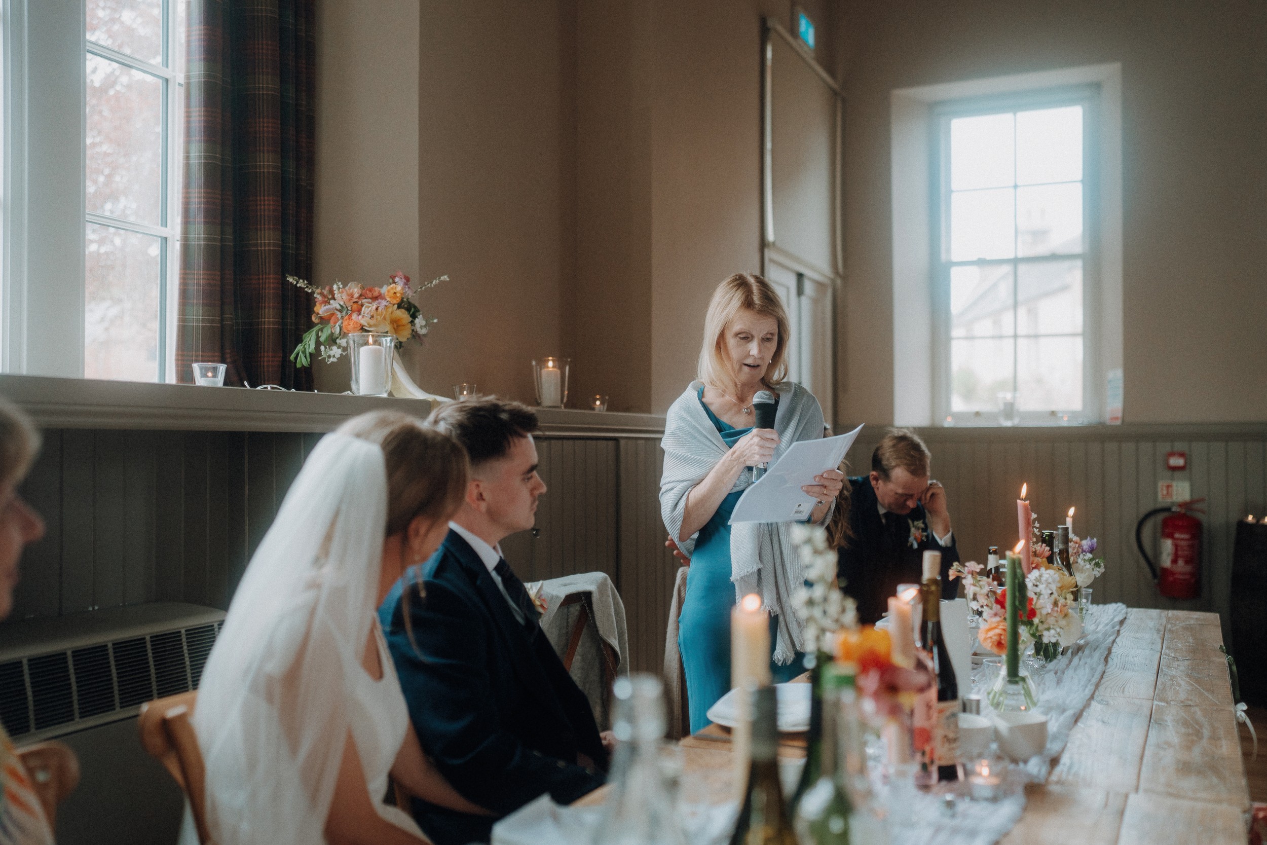 A woman stands and reads from a paper, giving a speech at a wedding reception while guests, including the bride and groom, listen at a decorated table.