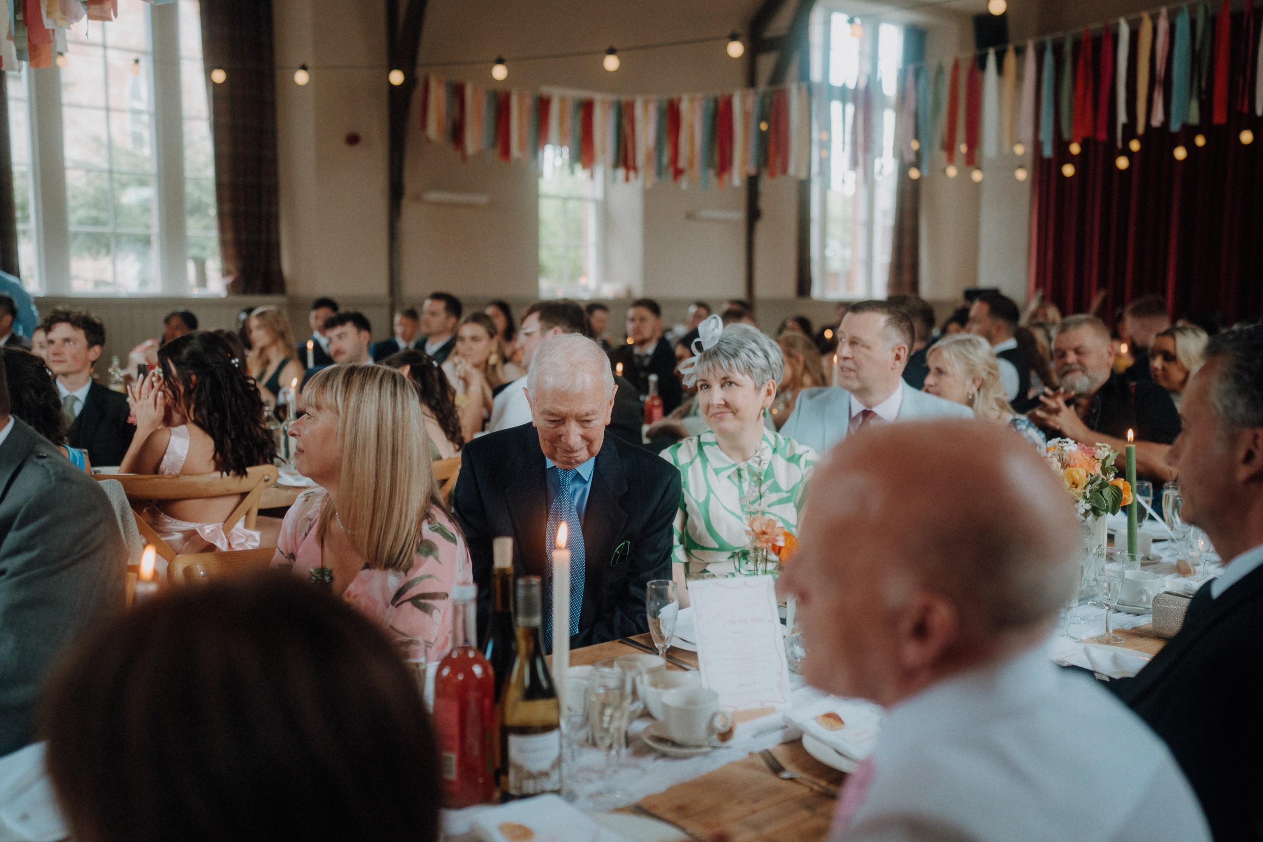 Guests seated at decorated tables in a well-lit hall, listening attentively during a formal event or celebration.