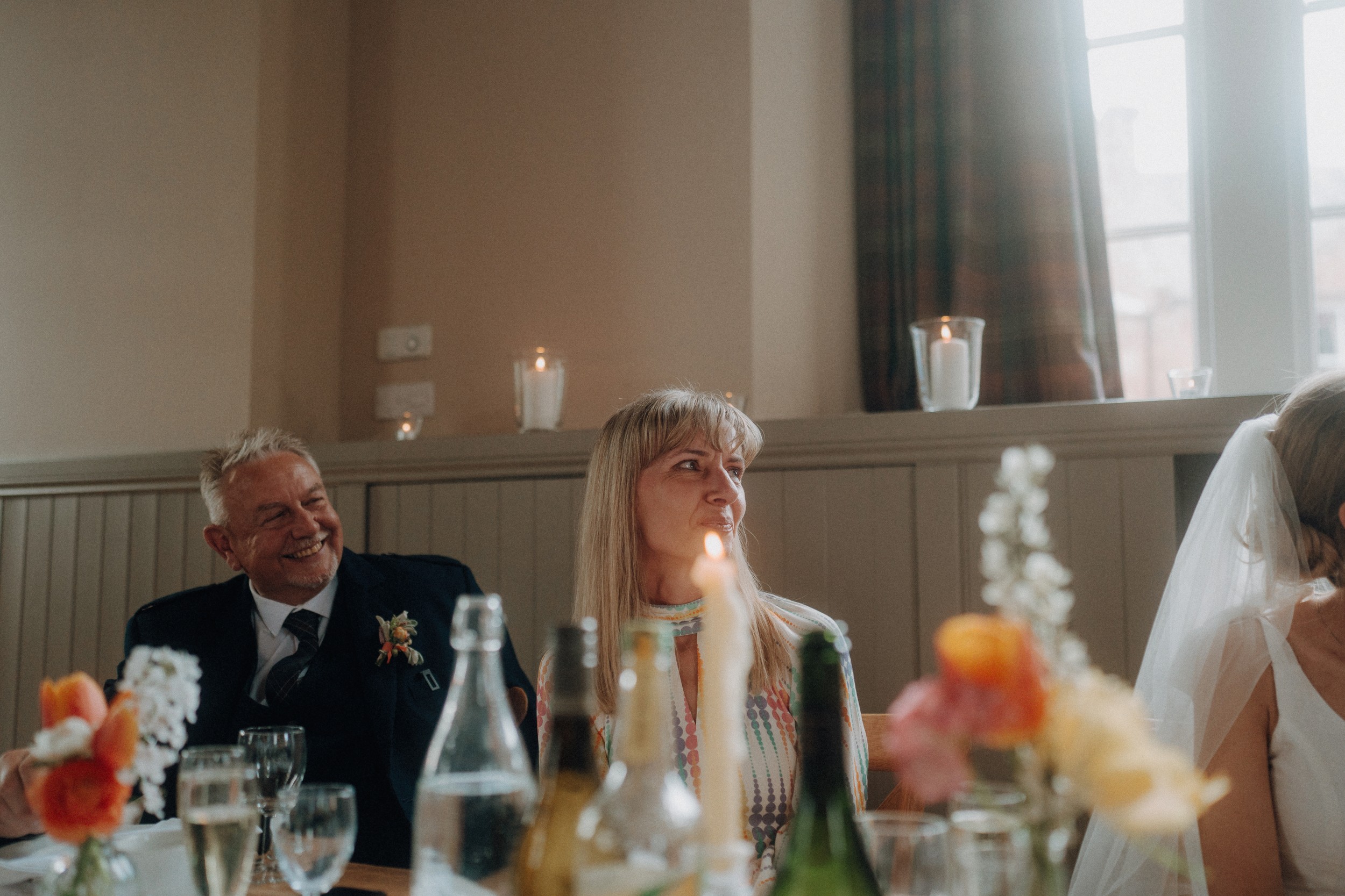 Two older adults sit at a decorated table with drinks and flowers, smiling and looking toward a bride in a white veil, inside a softly lit room with candles on the windowsill.
