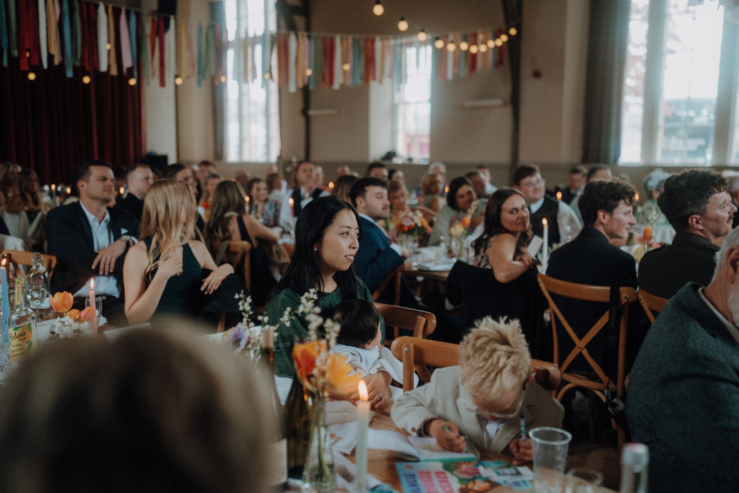 A group of people sit at decorated tables in a hall with colorful hanging ribbons, attending an event and listening attentively.