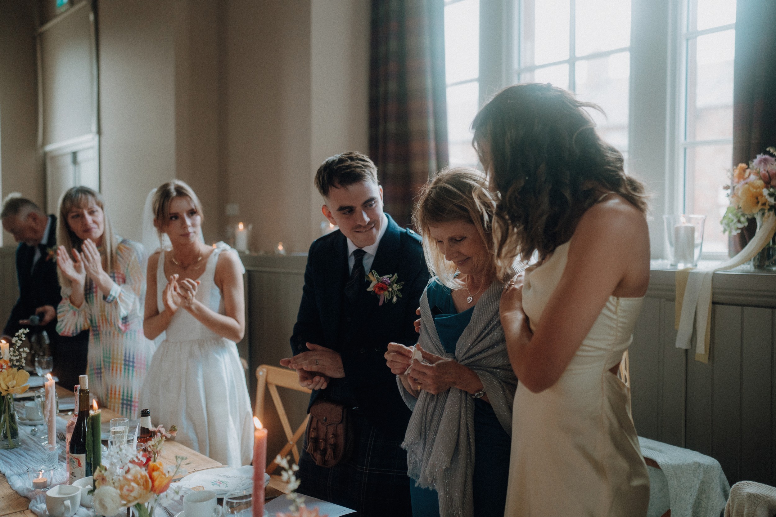 A group of people stand and sit around a decorated table at a formal event, with some clapping and others looking at a phone.