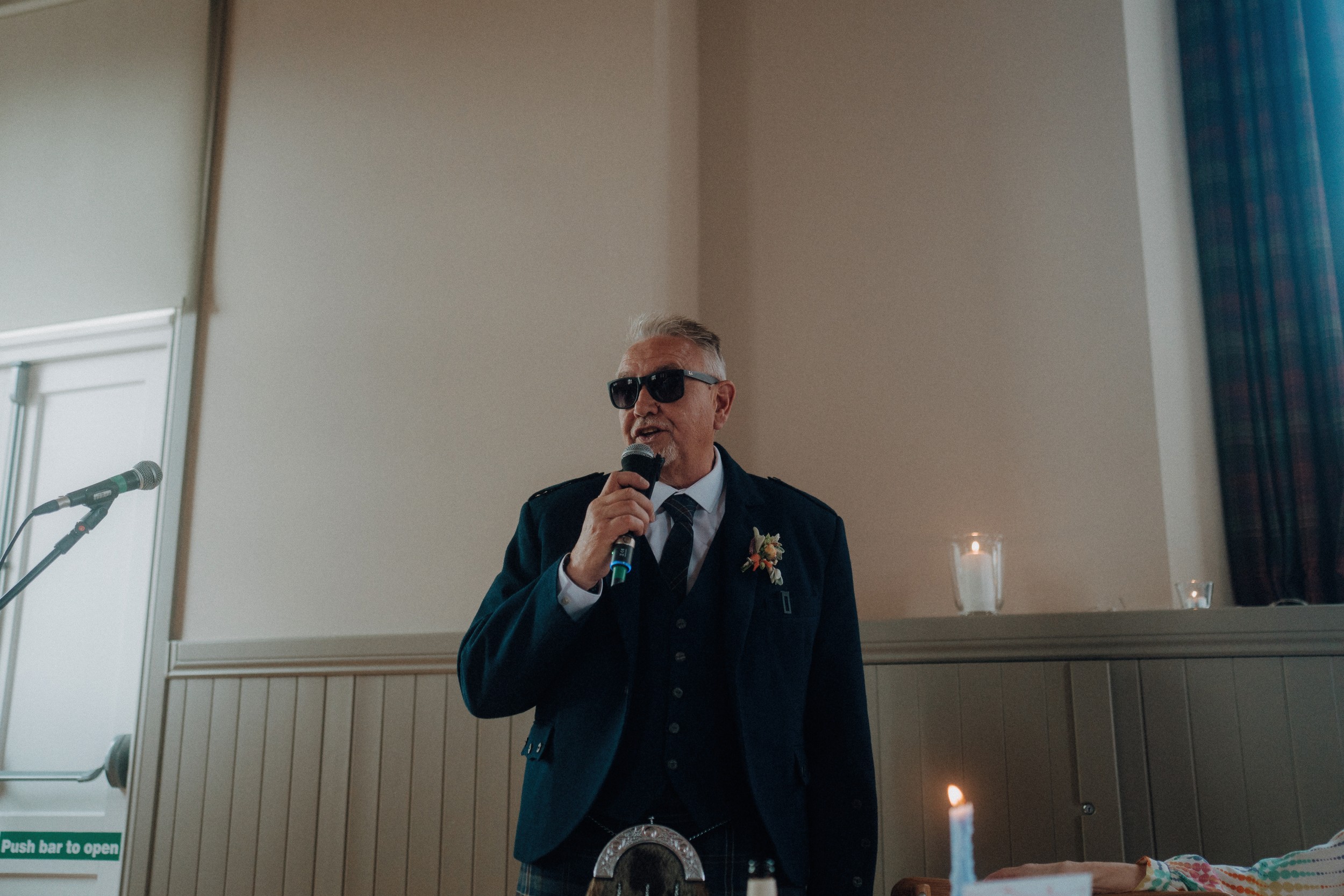 An older man in a suit and sunglasses stands indoors, speaking into a microphone. A lit candle and microphone stand are visible nearby.