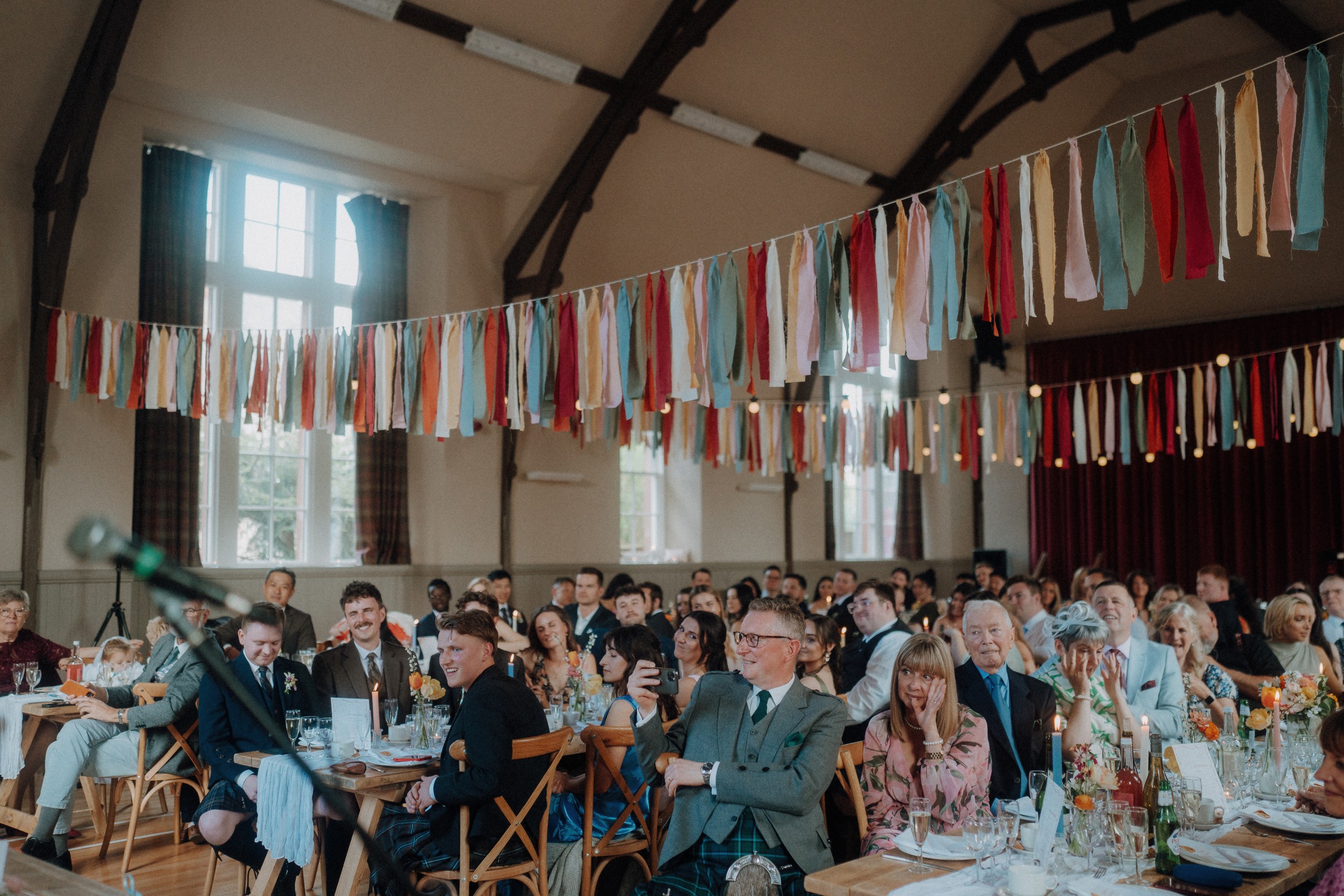 A group of people sit at decorated tables in a hall with high ceilings and colorful hanging ribbons, watching something out of frame.