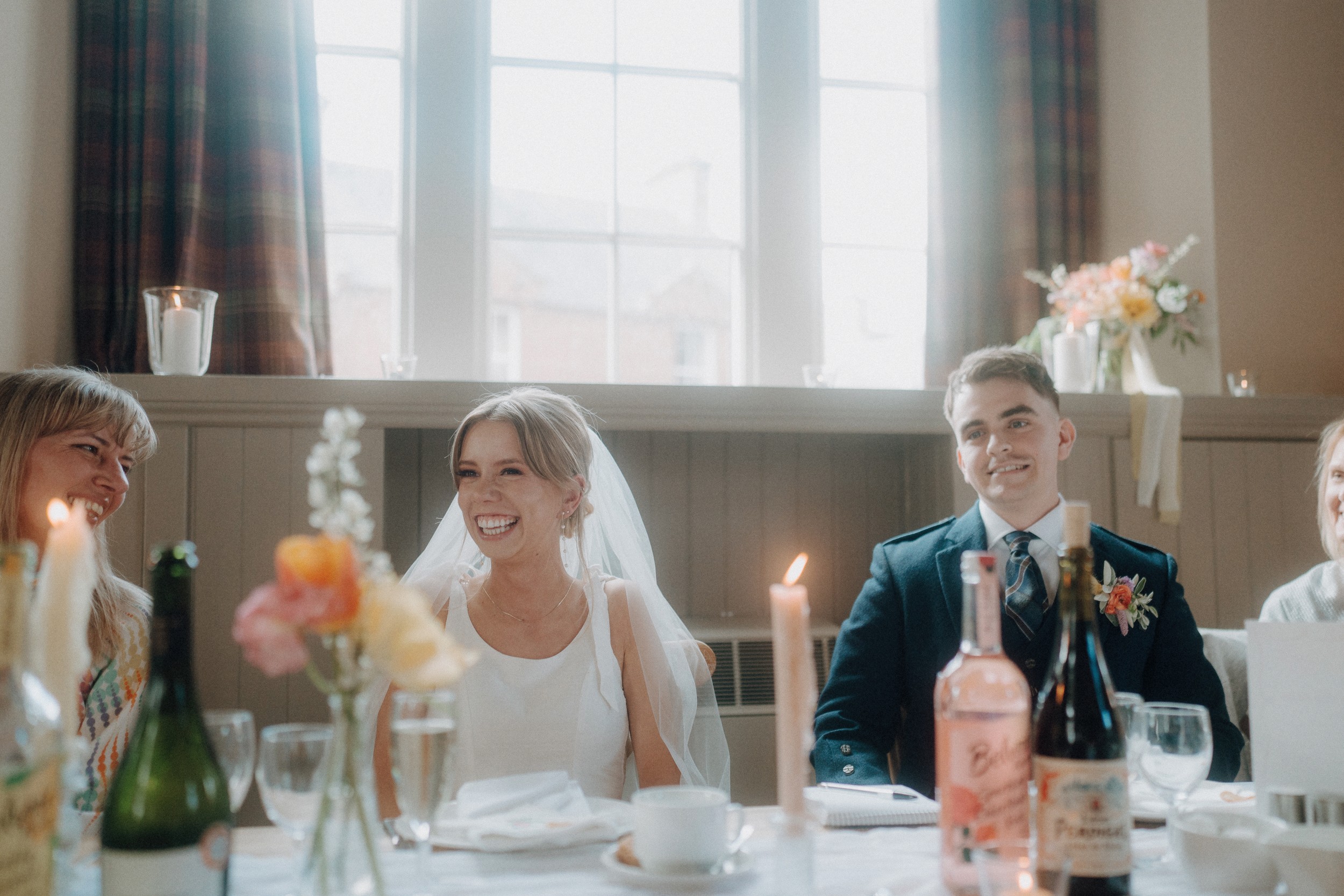 A bride and groom sit at a decorated table, smiling and surrounded by guests, candles, and flowers with daylight coming through large windows behind them.