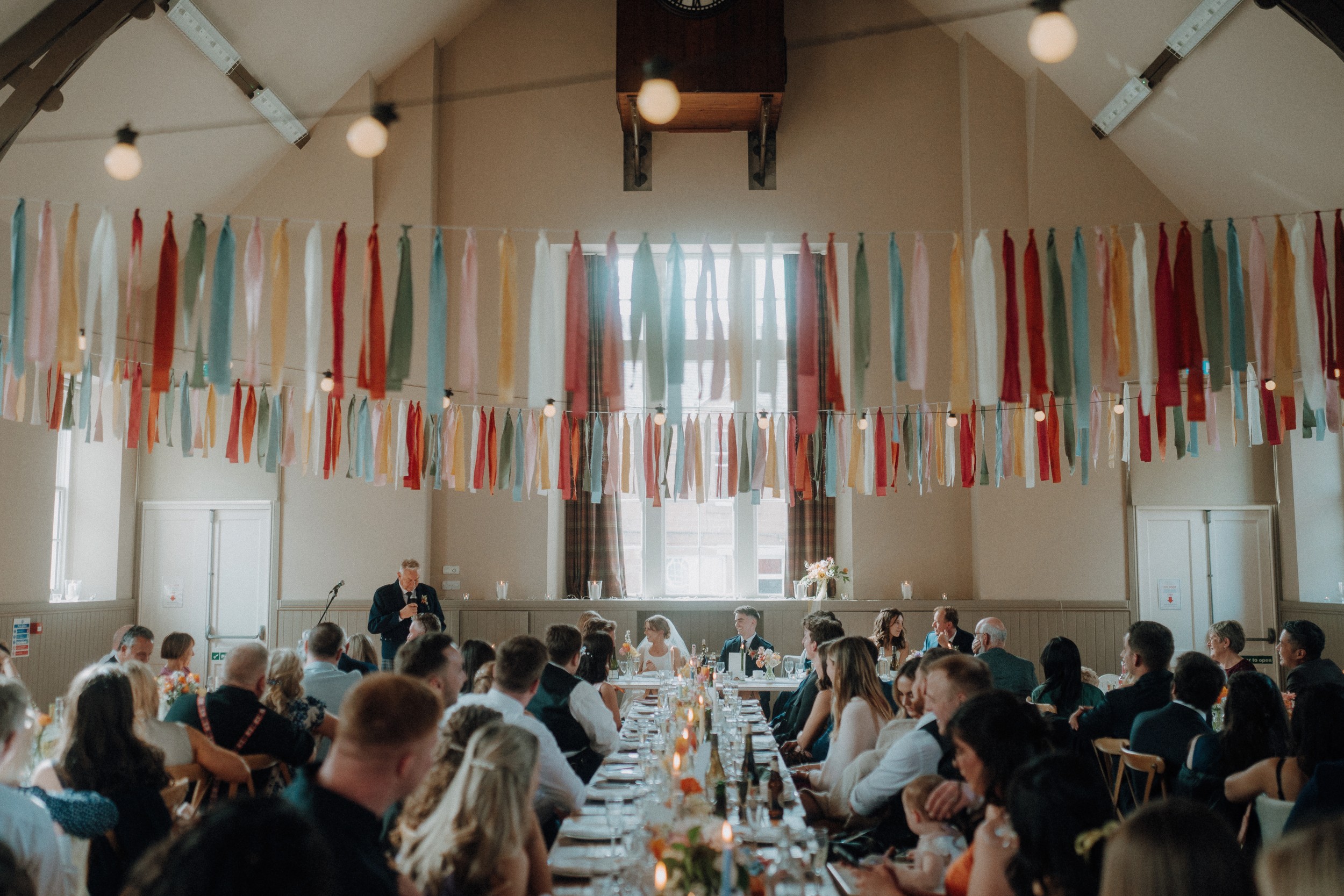 A large group of people seated at long tables in a decorated hall with colorful ribbons hanging from the ceiling, attending a formal event or celebration.