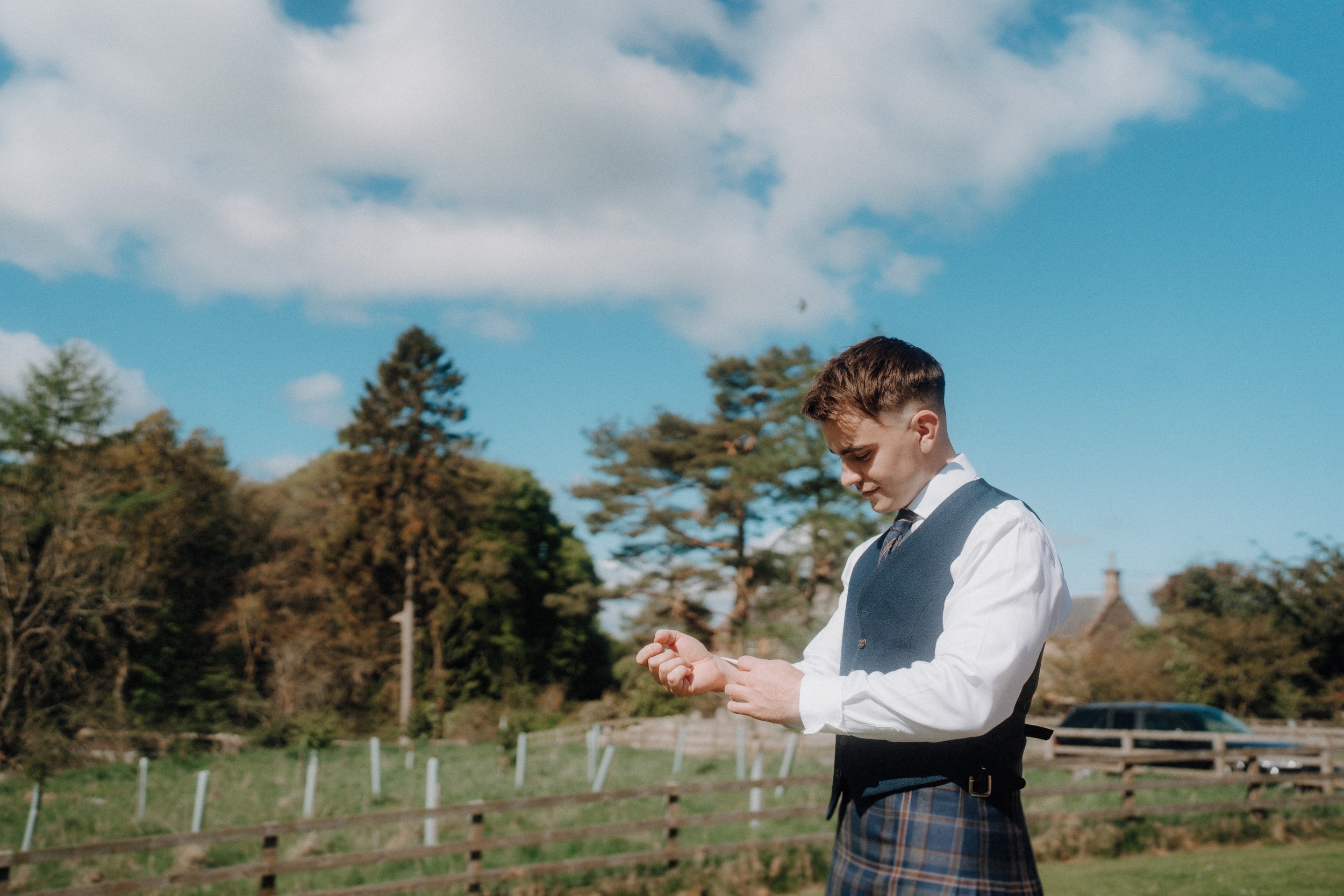 A man wearing a vest and tartan kilt adjusts his sleeve outdoors on a sunny day, with trees, a wooden fence, and a parked car in the background.