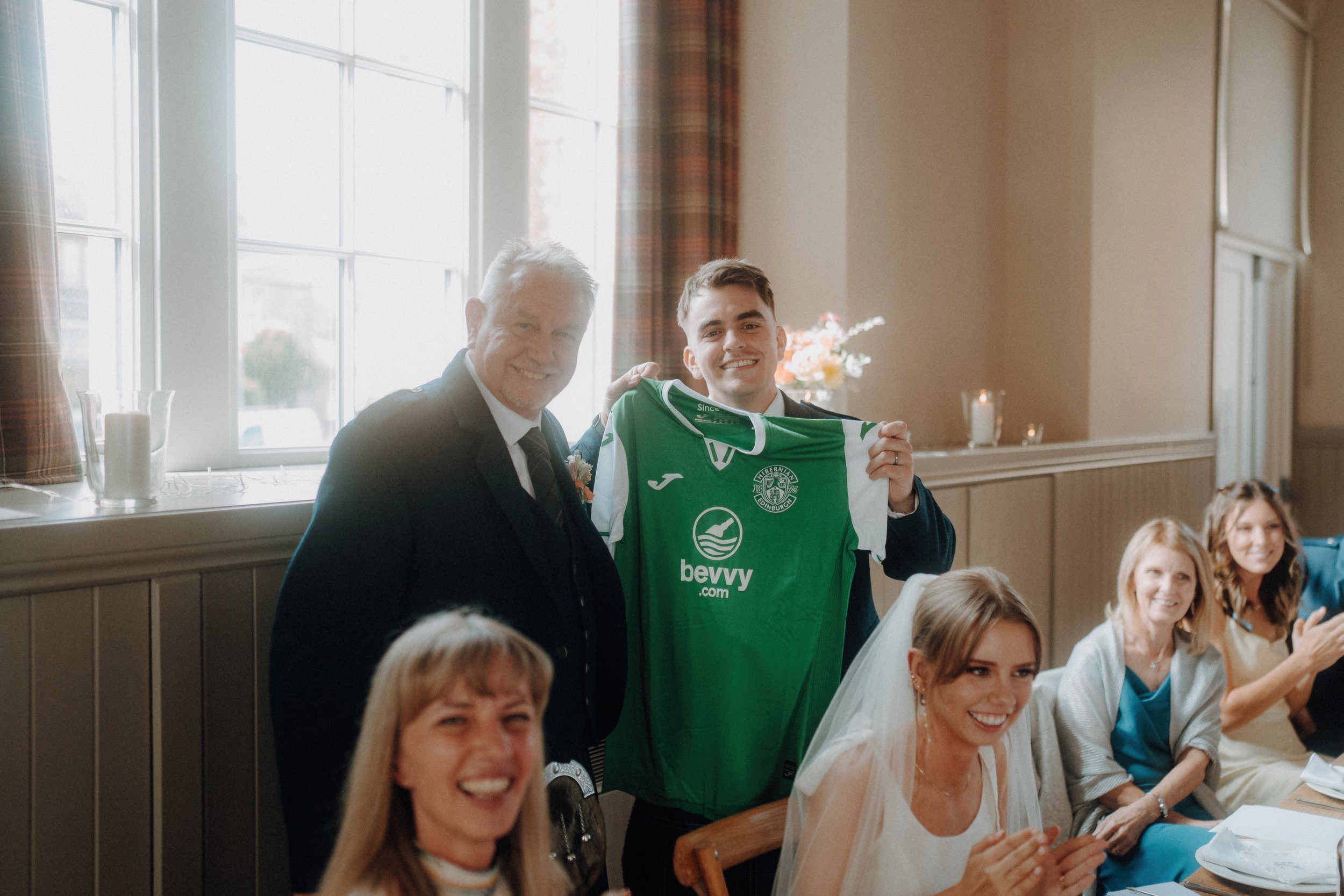 A young man holds up a green soccer jersey next to an older man, while several people seated at a table smile and clap in a well-lit room.
