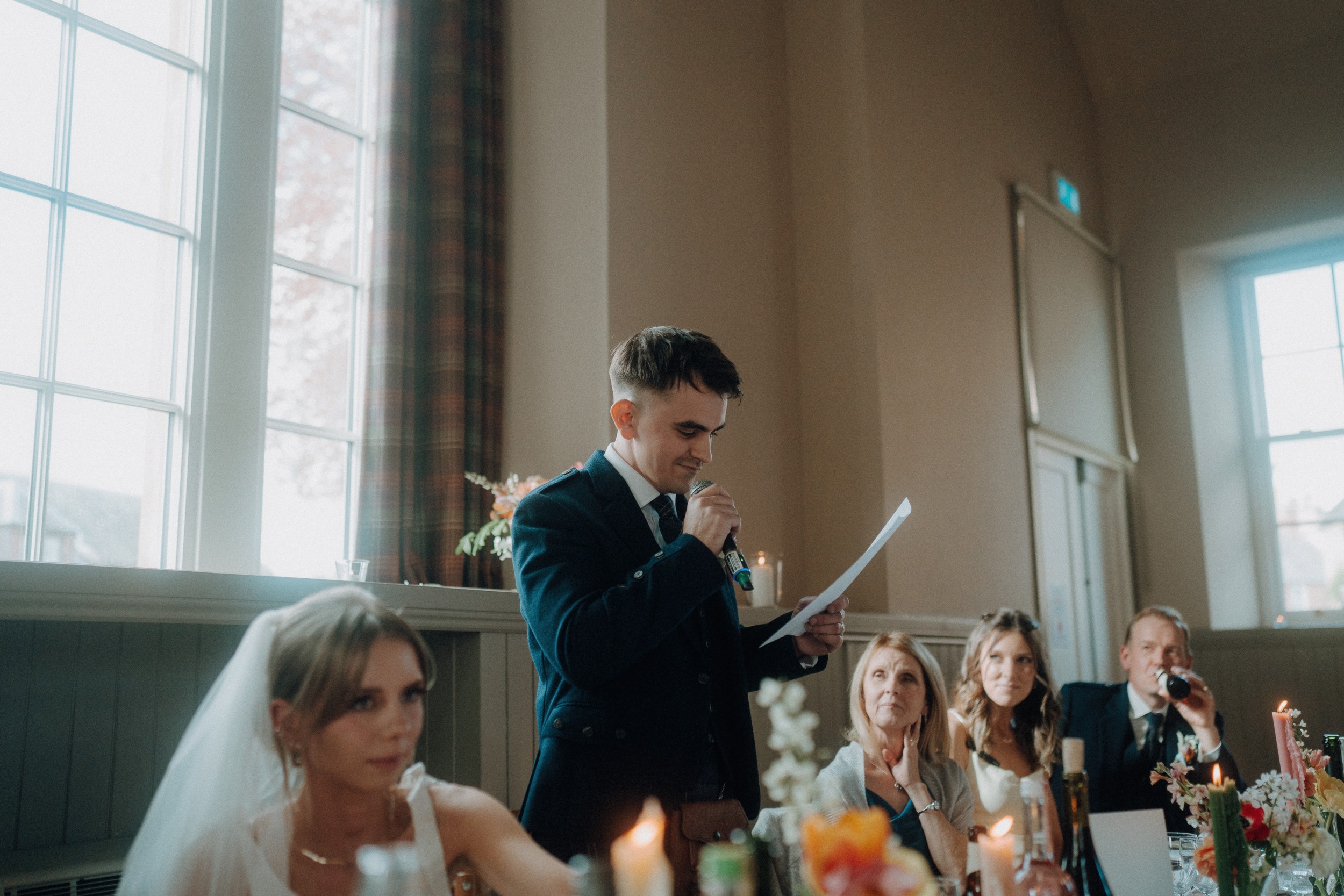 A man in a suit stands at a table reading from a paper, giving a speech at a wedding reception, with guests seated and listening.