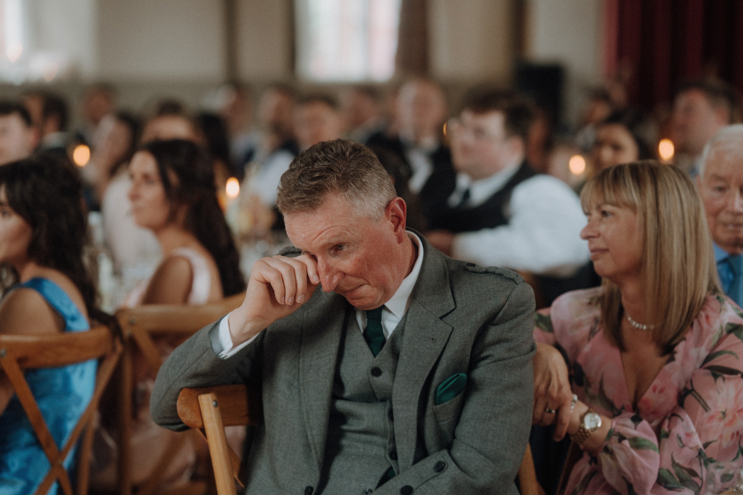 Man in a grey suit wipes his eye while seated at an event, surrounded by other people who are also seated and watching attentively.