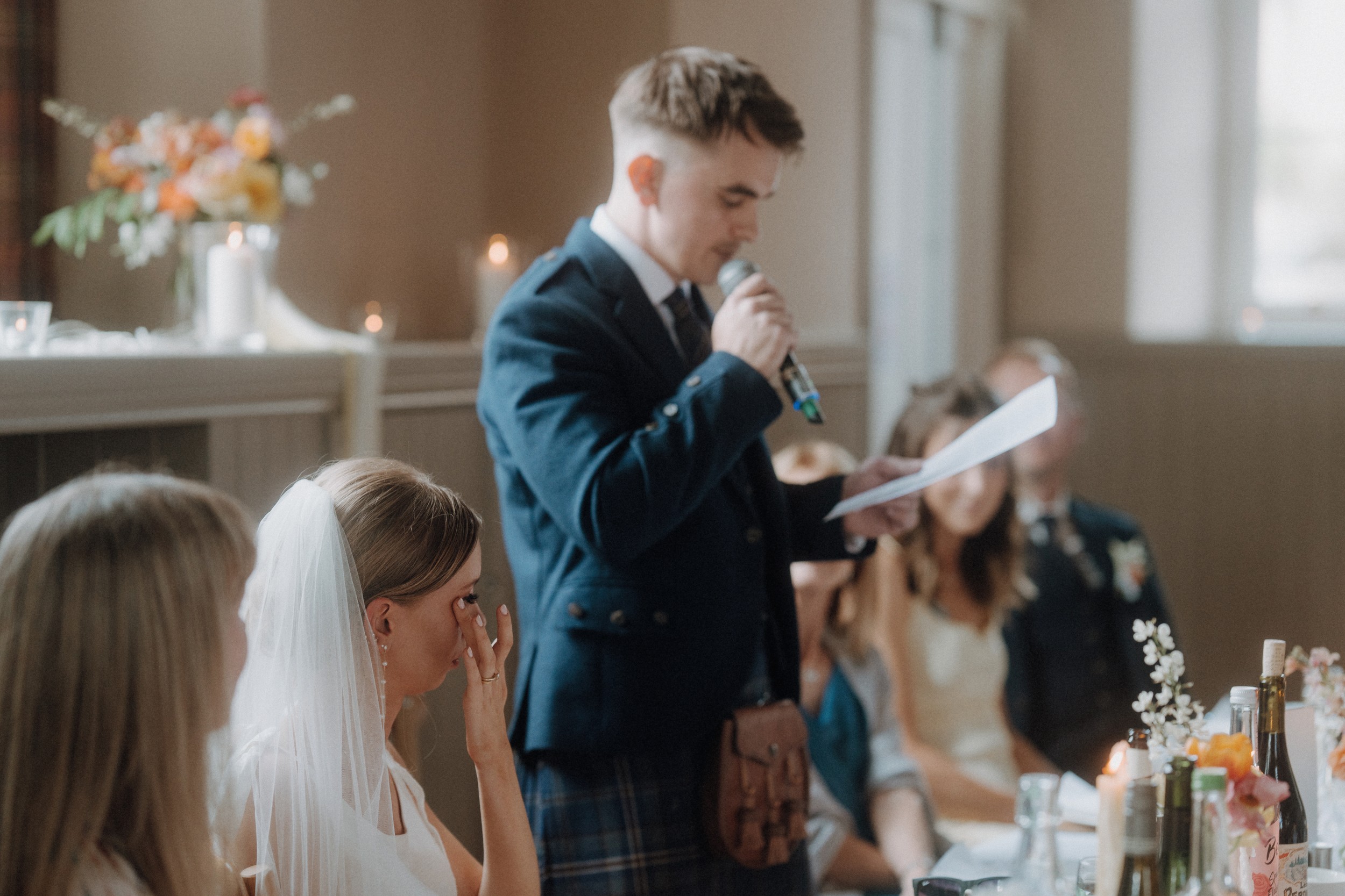 A man in formal attire gives a speech with a microphone at a wedding reception while the bride wipes her eyes; guests are seated at the table.