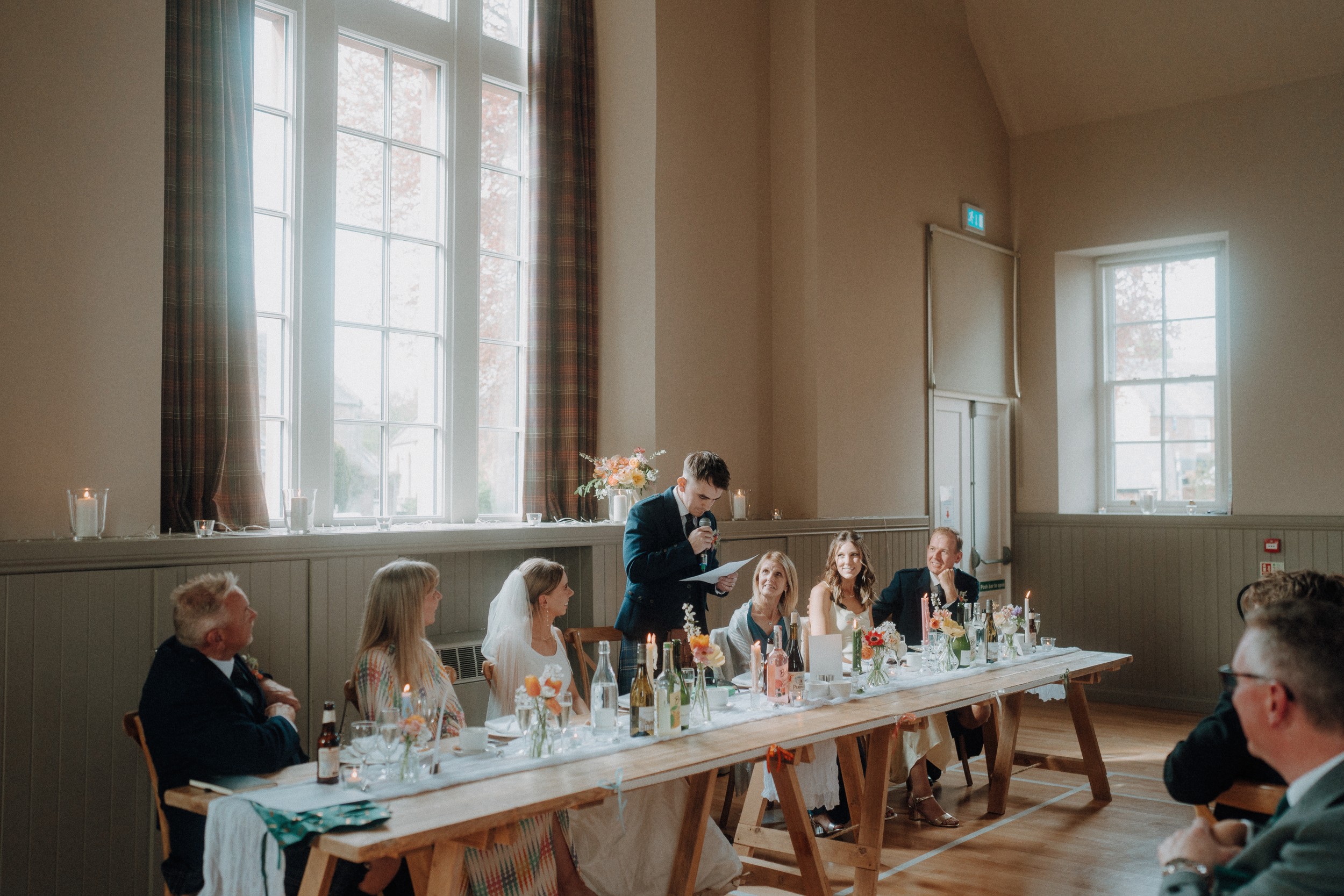 A man in a suit stands and speaks into a microphone at a long table during a formal event, with guests seated and listening in a bright, airy room.
